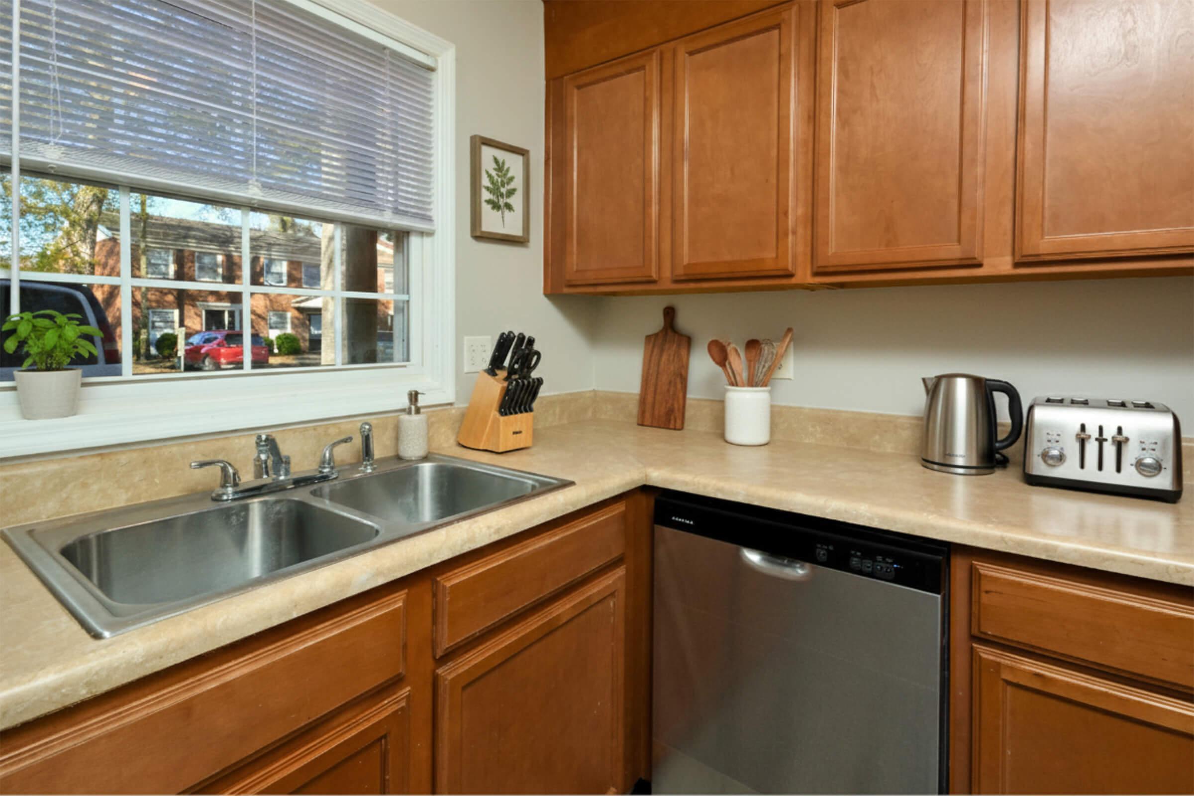 A modern kitchen featuring wooden cabinets, a double sink, a stainless steel dishwasher, a countertop with a kettle and toaster, and a wooden cutting board. A small plant is on the windowsill, and natural light is streaming in through a window with blinds.