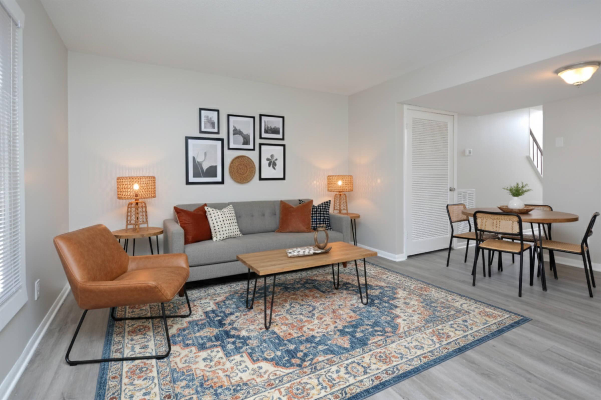 A modern living room with a gray sofa adorned with decorative pillows, two stylish lamps on either side, and a patterned area rug. A wooden coffee table sits in the center, surrounded by an inviting armchair. In the background, there's a dining area with a small table and chairs, enhancing the cozy ambiance.