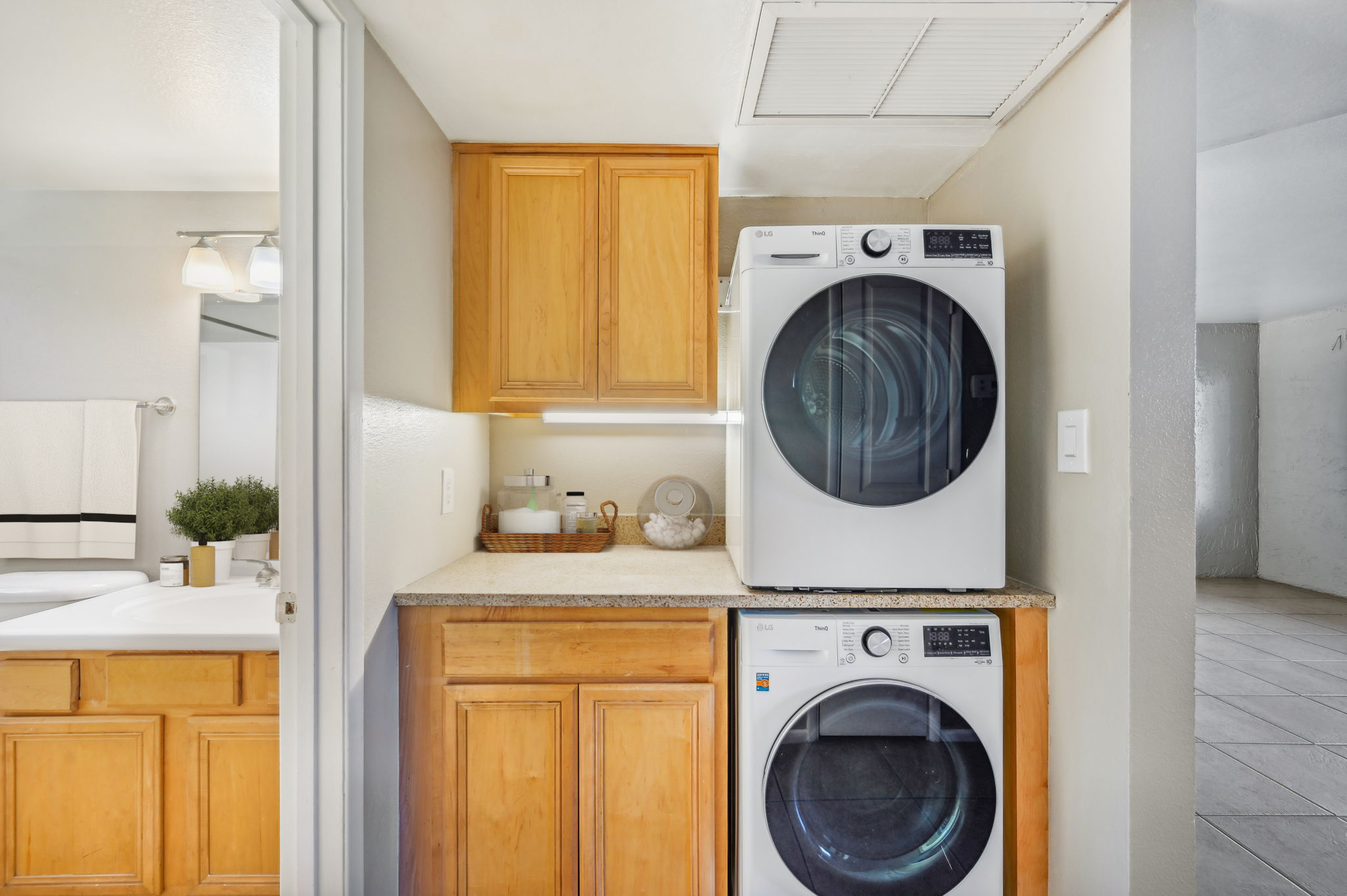 A compact laundry area featuring a stacked washer and dryer in white, situated in a well-lit room with light-colored walls and wooden cabinetry. The countertop displays a decorative basket and various laundry supplies. A bathroom can be glimpsed through an open door in the background.