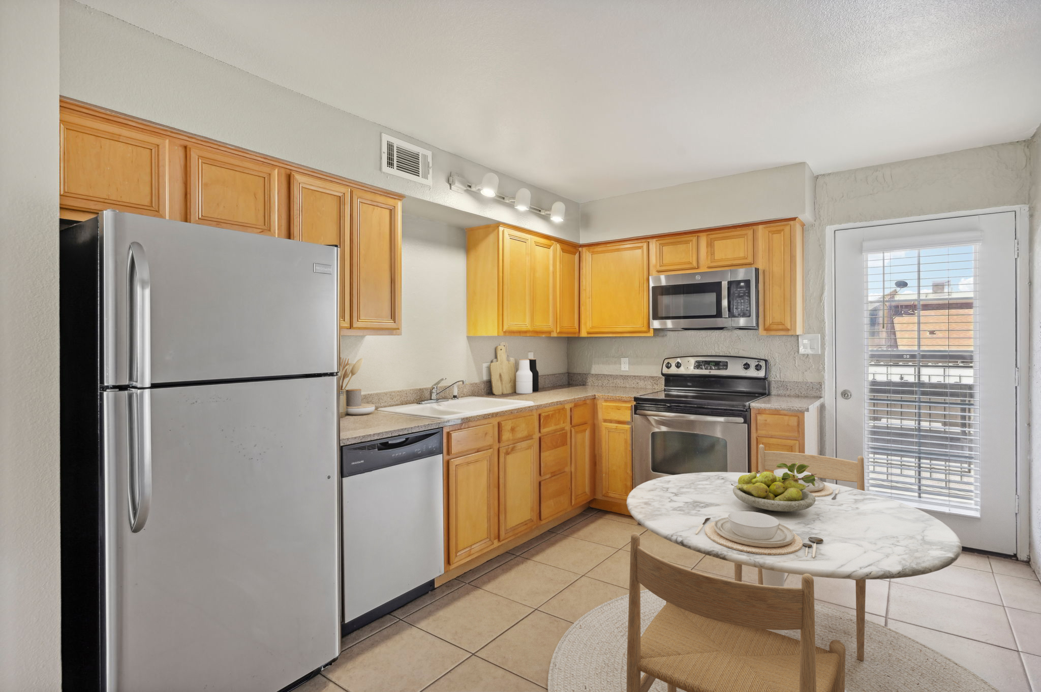 Modern kitchen featuring light wood cabinets, stainless steel appliances including a refrigerator, dishwasher, and microwave. A round marble table with a bowl of green apples sits in the foreground, beside a sliding glass door leading to an outdoor area. Neutral-colored tile flooring complements the space.