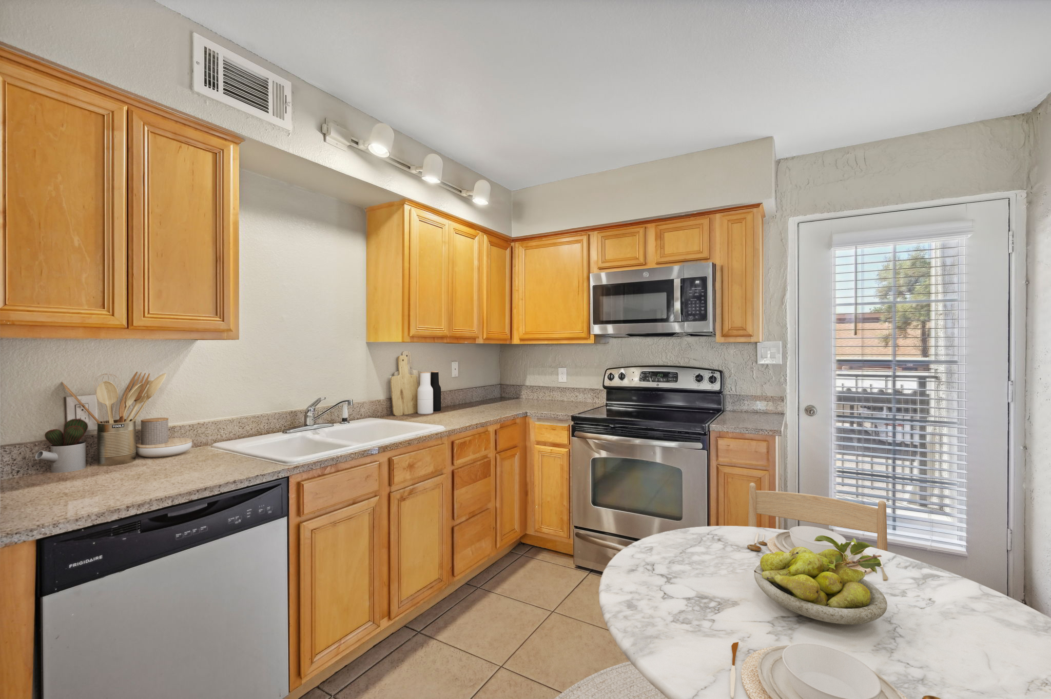A modern kitchen featuring wooden cabinets, a stainless steel microwave and oven, and a dishwasher. The countertop is made of light-colored stone, with a sink and kitchen utensils displayed. There is a round table with a marble top and a bowl of green fruits, along with a door leading to a patio with sunlight filtering in.