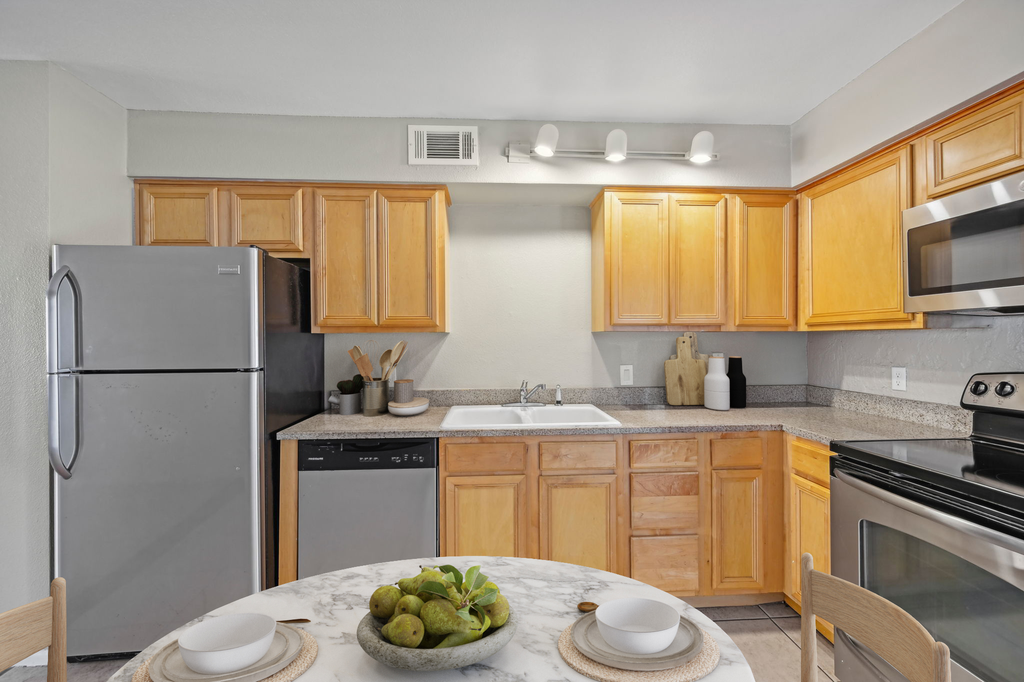 A modern kitchen featuring wooden cabinets, a stainless steel refrigerator, a dishwasher, and a stainless steel oven. The countertop is made of granite and includes a sink. A round table with a marble top is set for dining, showcasing a bowl of green fruit, with cutlery and placemats visible.