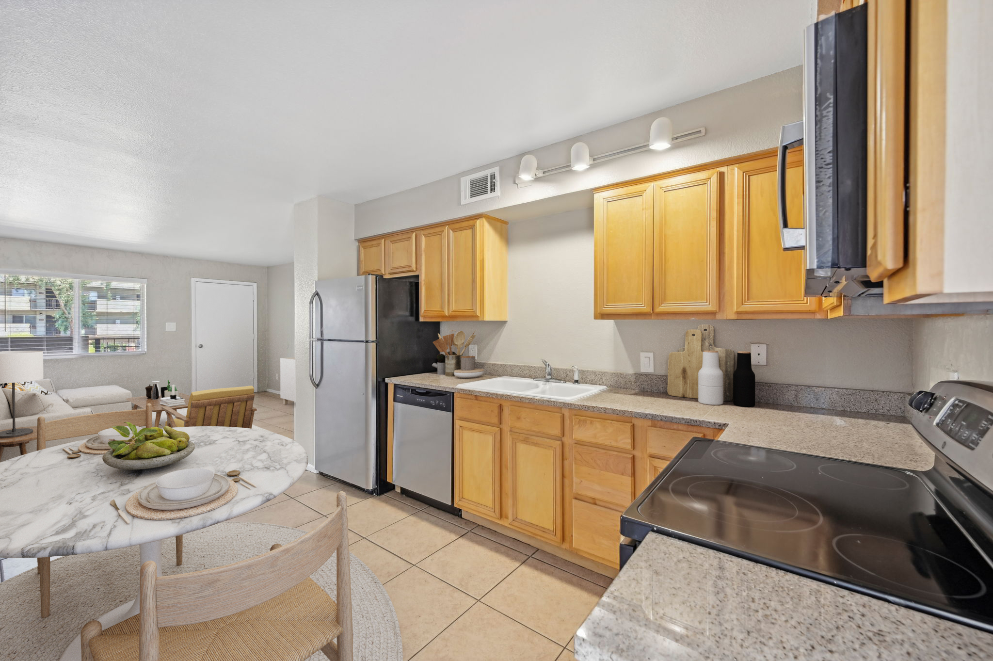A modern kitchen featuring light wood cabinetry, a marble dining table with fruit, stainless steel appliances including a refrigerator, and a stove. The space has beige tiled flooring and soft lighting, with a living area visible in the background.