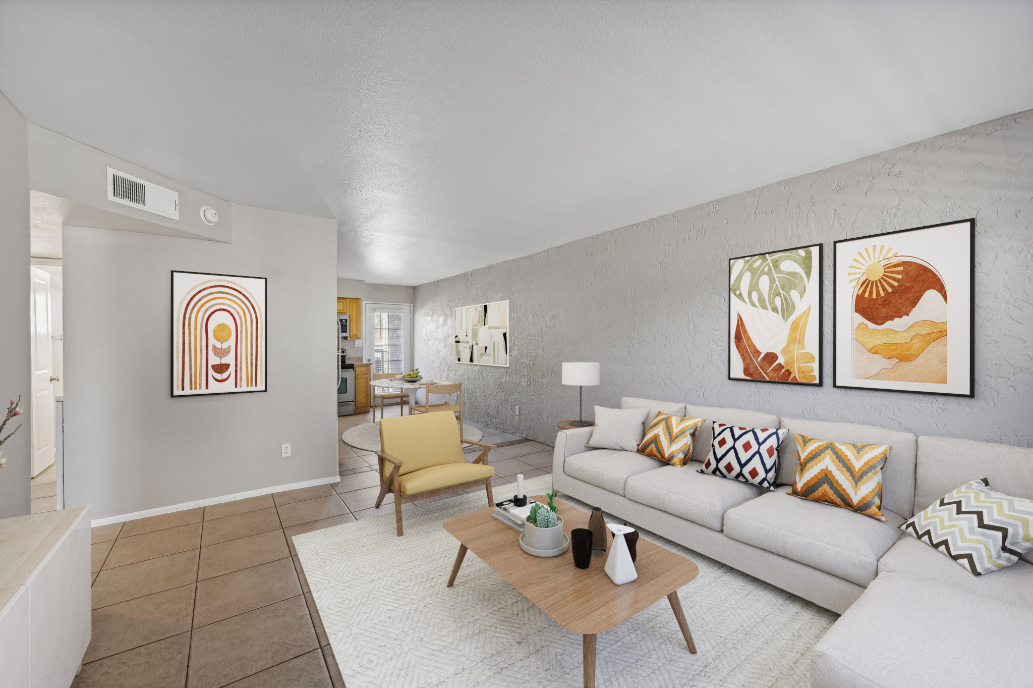 A modern living room featuring a light-colored sofa adorned with vibrant patterned cushions. A wooden coffee table sits in front, with decorative items on it. The walls are painted gray, and two framed artworks hang above the sofa. To the left, a glimpse of a dining area and kitchen is visible, enhancing the open layout.