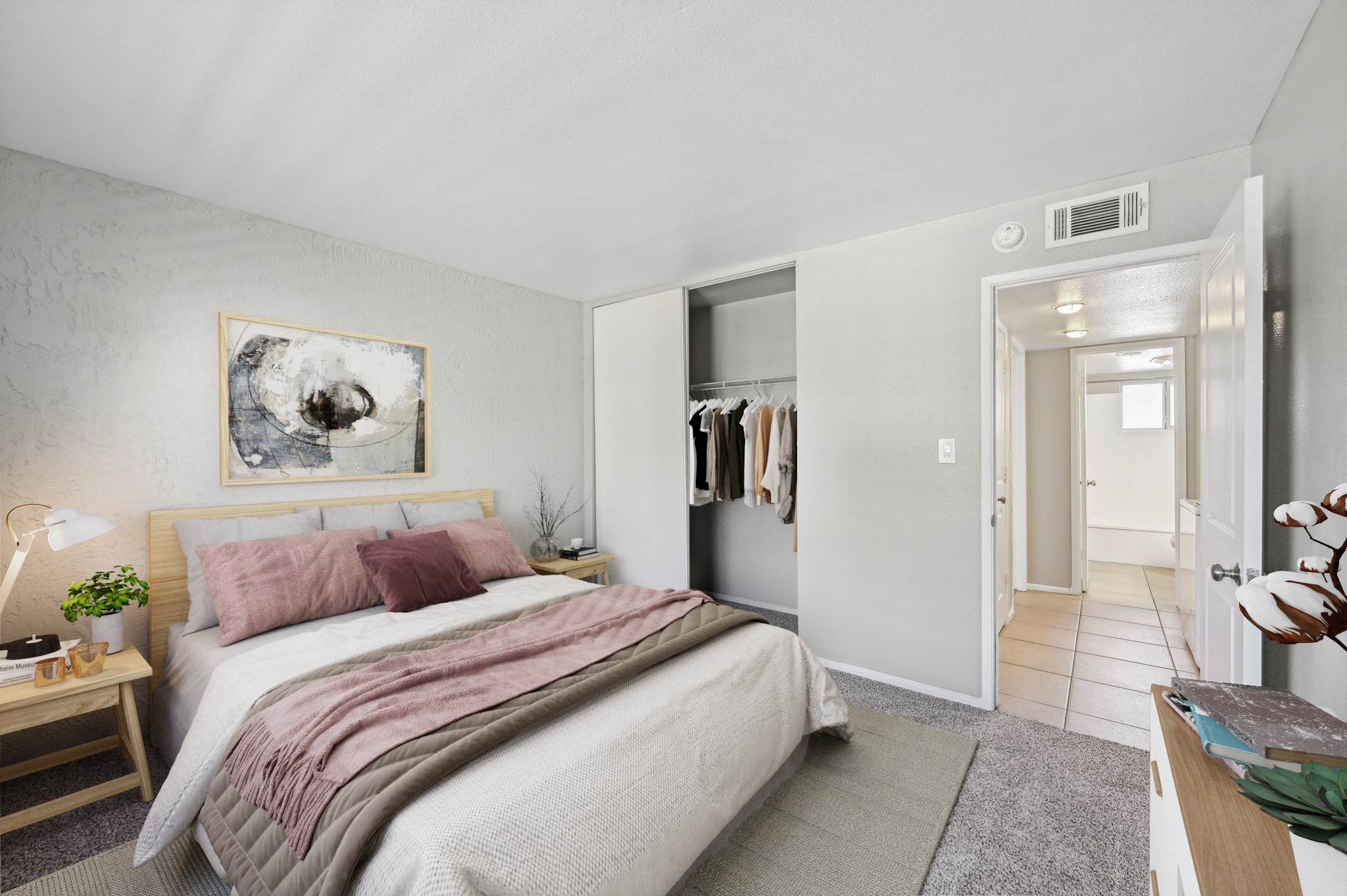 A well-decorated bedroom featuring a cozy bed with pink and neutral bedding, a nightstand with a lamp and a potted plant, and a closet with hanging clothes. The walls are painted light gray, and there's a piece of abstract artwork above the bed. The room is bright and inviting, with a door leading to another space.
