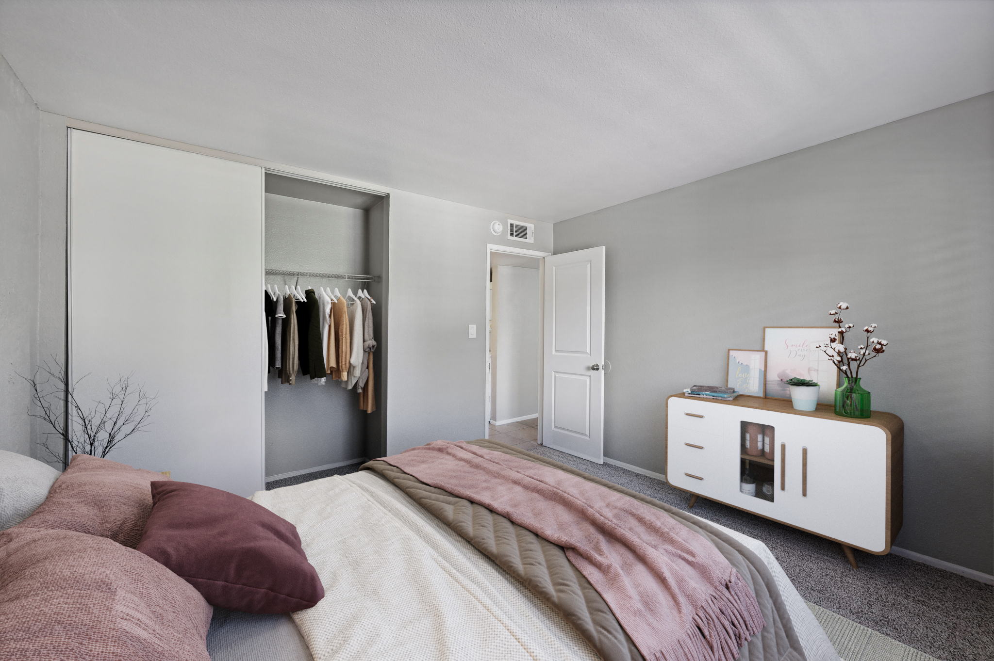 A modern bedroom featuring a neatly made bed with pillows and a cozy blanket. A wardrobe with hanging clothes is visible, along with a white dresser adorned with decor items. Soft gray walls and natural light enhance the inviting atmosphere of the room.