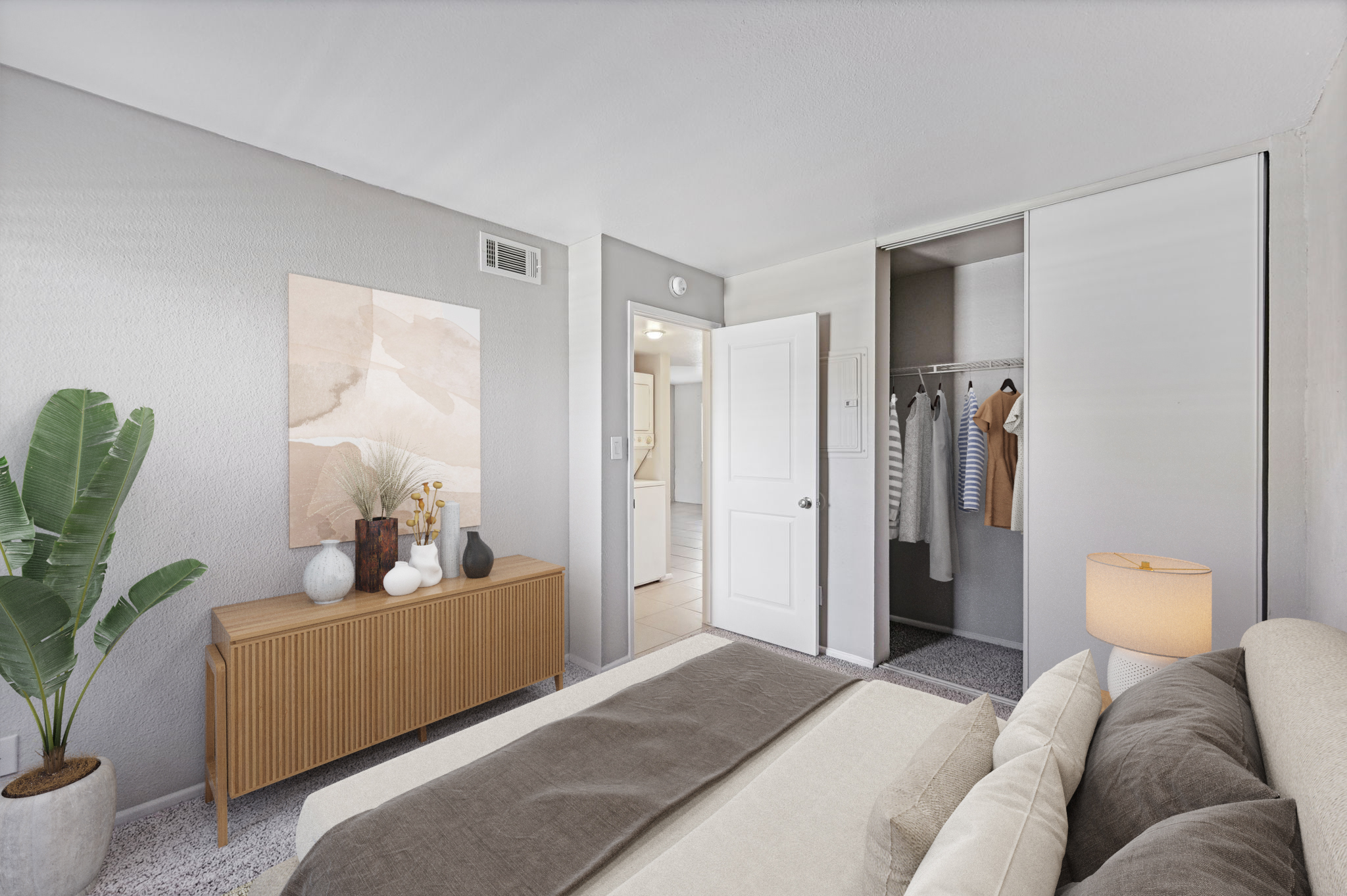 A cozy bedroom featuring a neatly made bed with beige linens, a light gray wall, and a wooden dresser. There is a potted plant in one corner, a modern lamp on the nightstand, and an open closet displaying hanging clothes. A soft art piece hangs on the wall above the dresser, contributing to the room's serene ambiance.