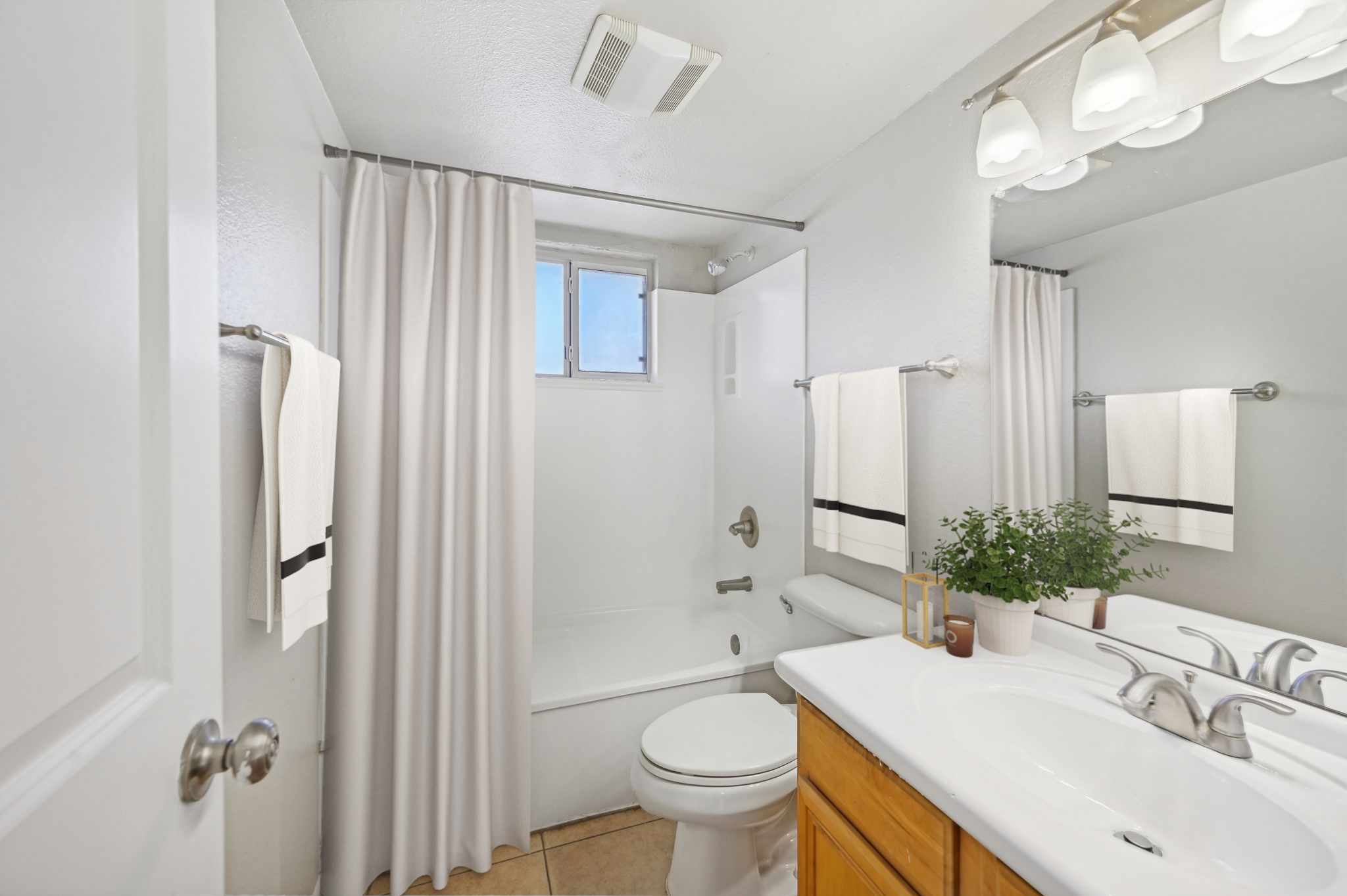 A modern bathroom featuring a white bathtub with a shower curtain, a sink with a double faucet, a large mirror, and light fixtures. The walls are painted light gray, and there are neatly hung towels. A small potted plant adds a touch of greenery to the countertop.