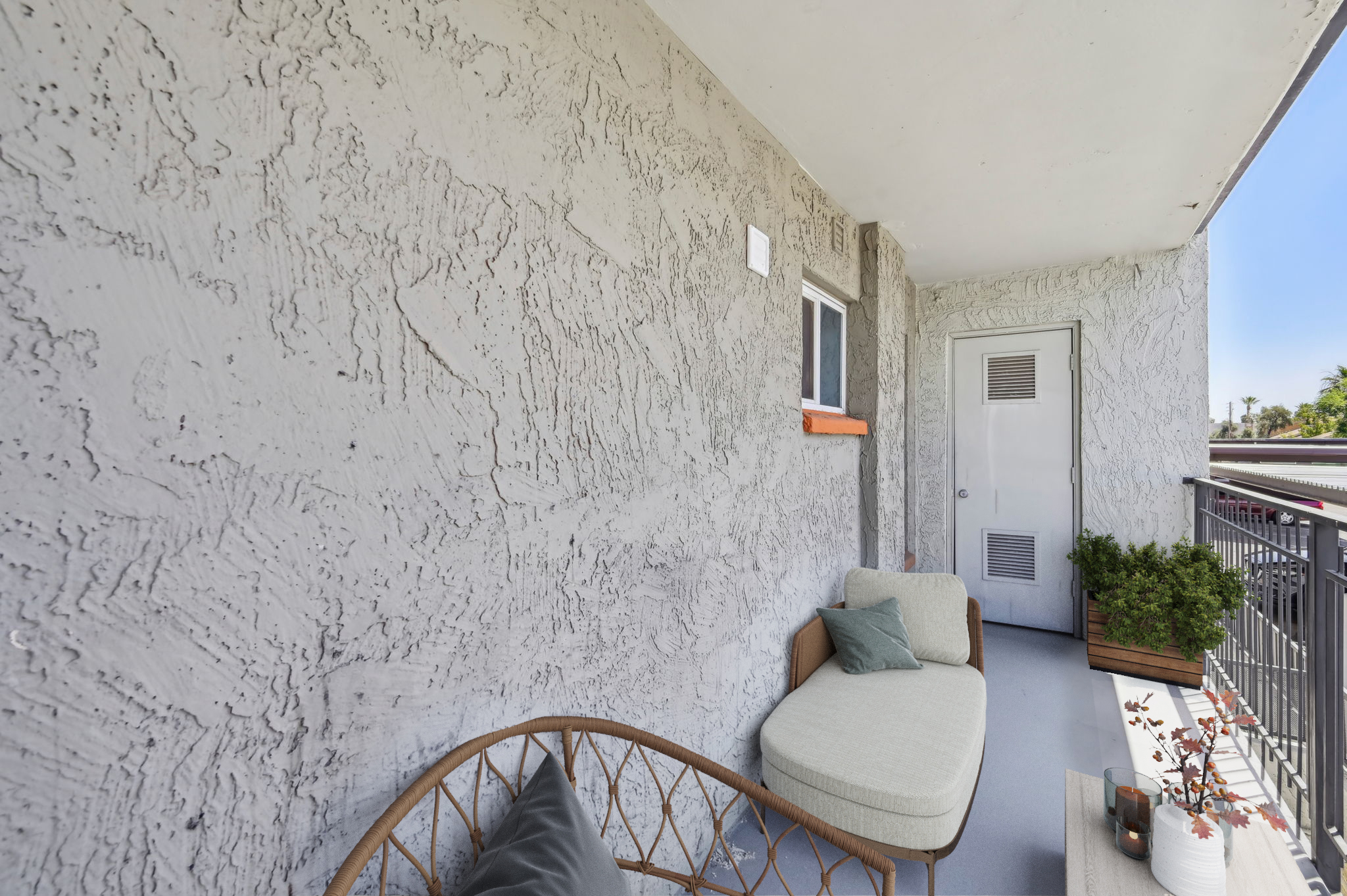 A simple balcony with textured gray walls. It features a curved wicker bench with a light cushion and a small potted plant, alongside a door that leads inside. The scene is well-lit and the balcony offers a view of the outdoors.