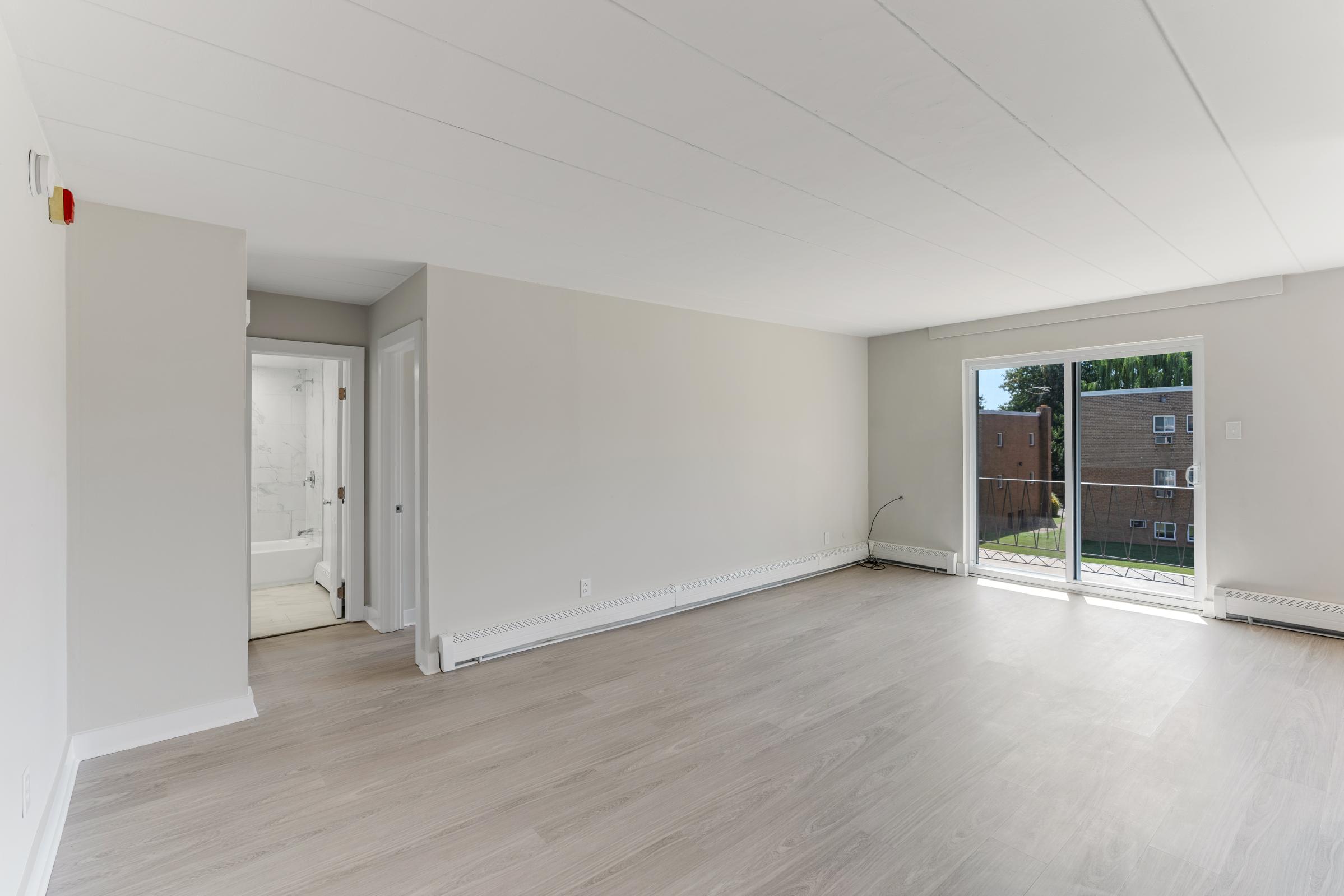 Spacious, modern living room with light-colored walls and new flooring. An open doorway leads to a bathroom, and sliding glass doors open to a balcony. The room is bright with natural light, offering a clean and inviting atmosphere.