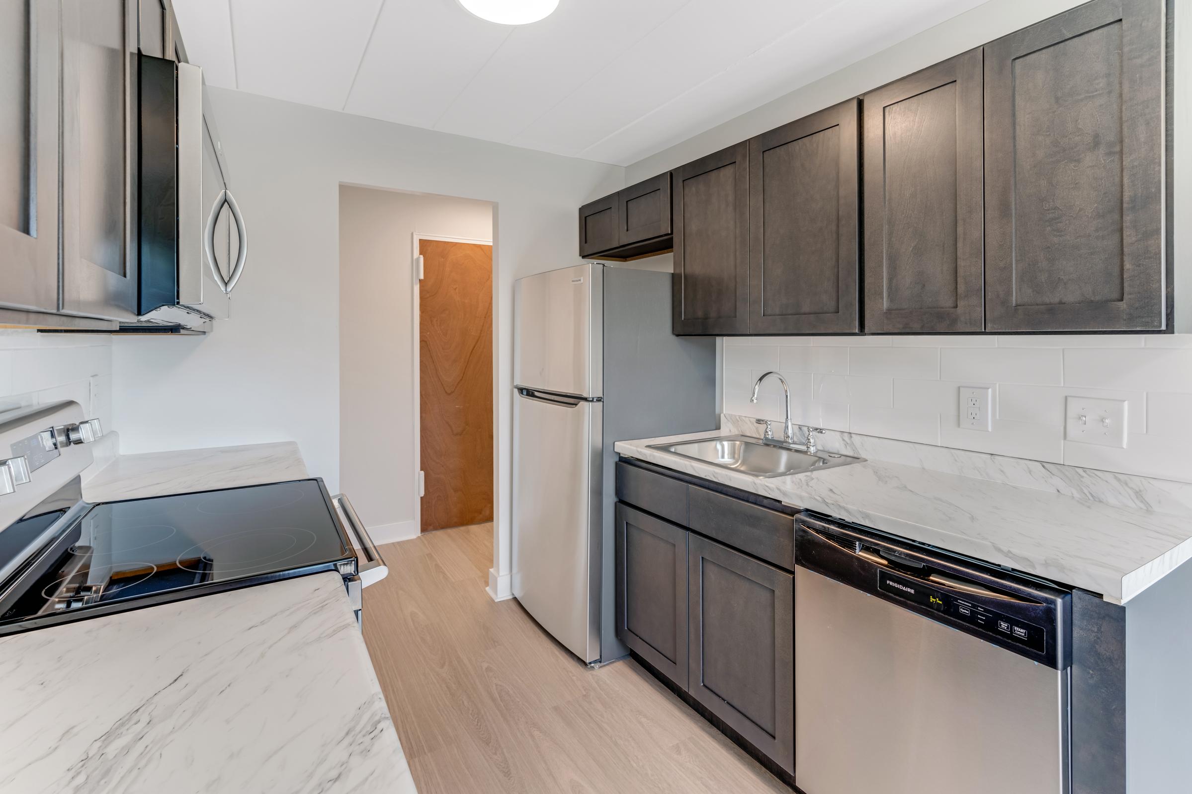 Modern kitchen featuring dark wood cabinets, stainless steel appliances including a refrigerator and dishwasher, and a sleek marble countertop. The space has an open layout with light-colored walls and flooring, providing a bright and inviting atmosphere.