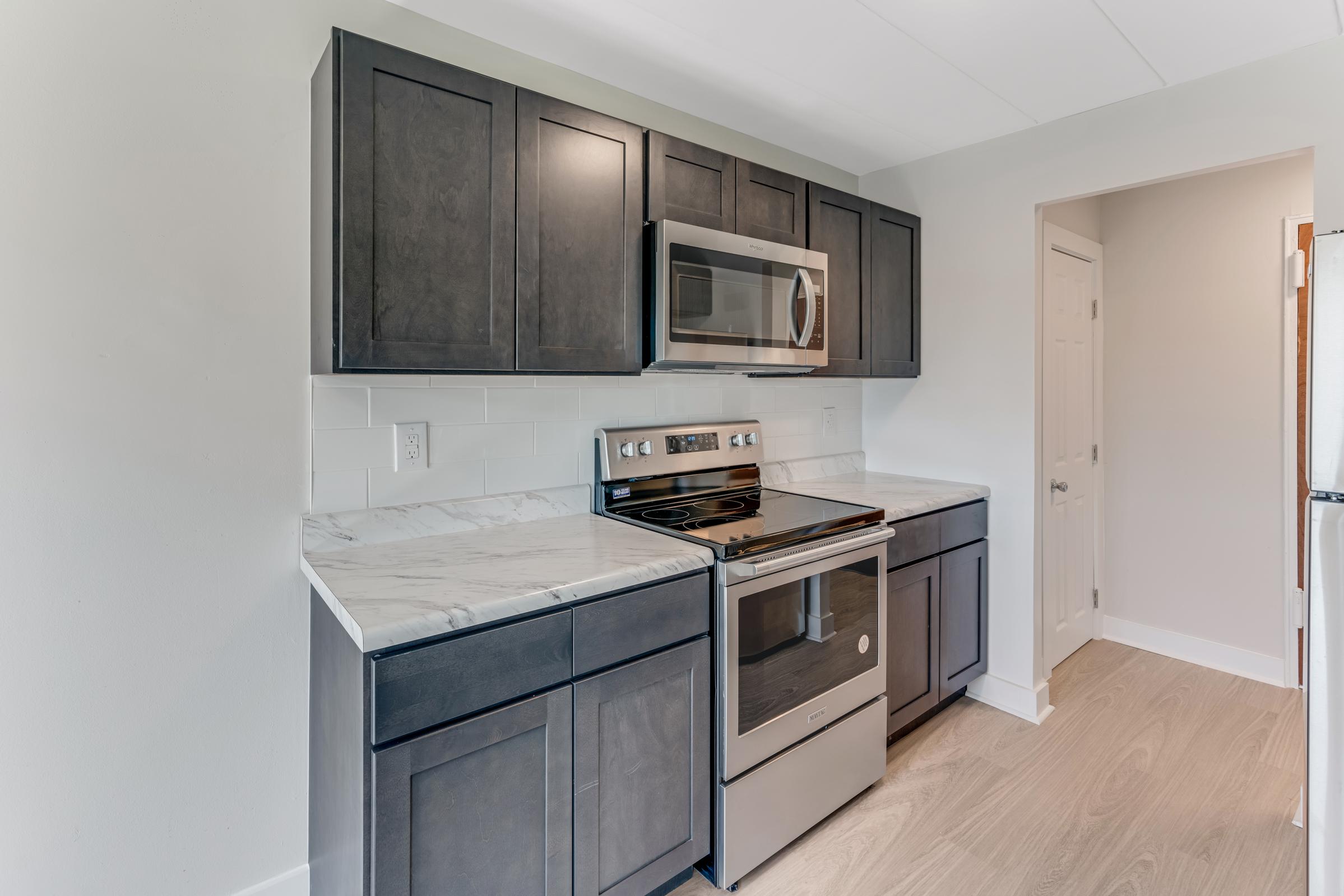 Modern kitchen featuring dark wood cabinetry, a stainless steel microwave, and an oven. The countertops are made of marble, and the flooring is light-colored wood. The walls are painted in a neutral tone, creating a bright and spacious feel. A door leading to another room is visible in the background.