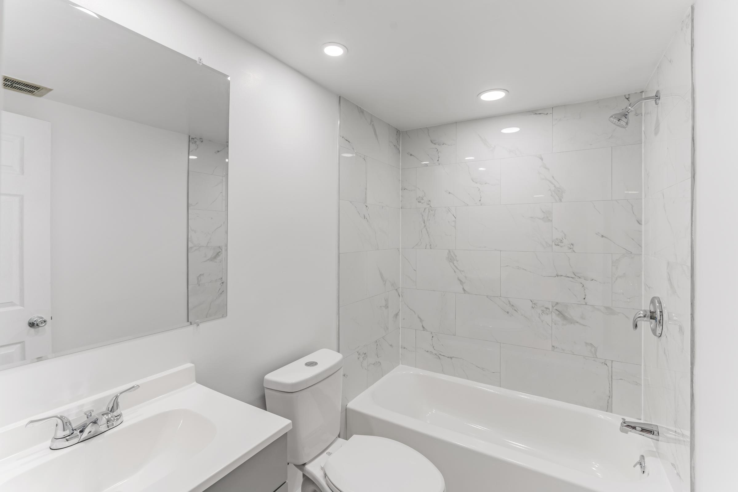 A modern bathroom featuring white marble walls, a bathtub with a shower, a sleek sink with a mirror above it, and a toilet. The lighting is bright, and the overall design is clean and minimalist.