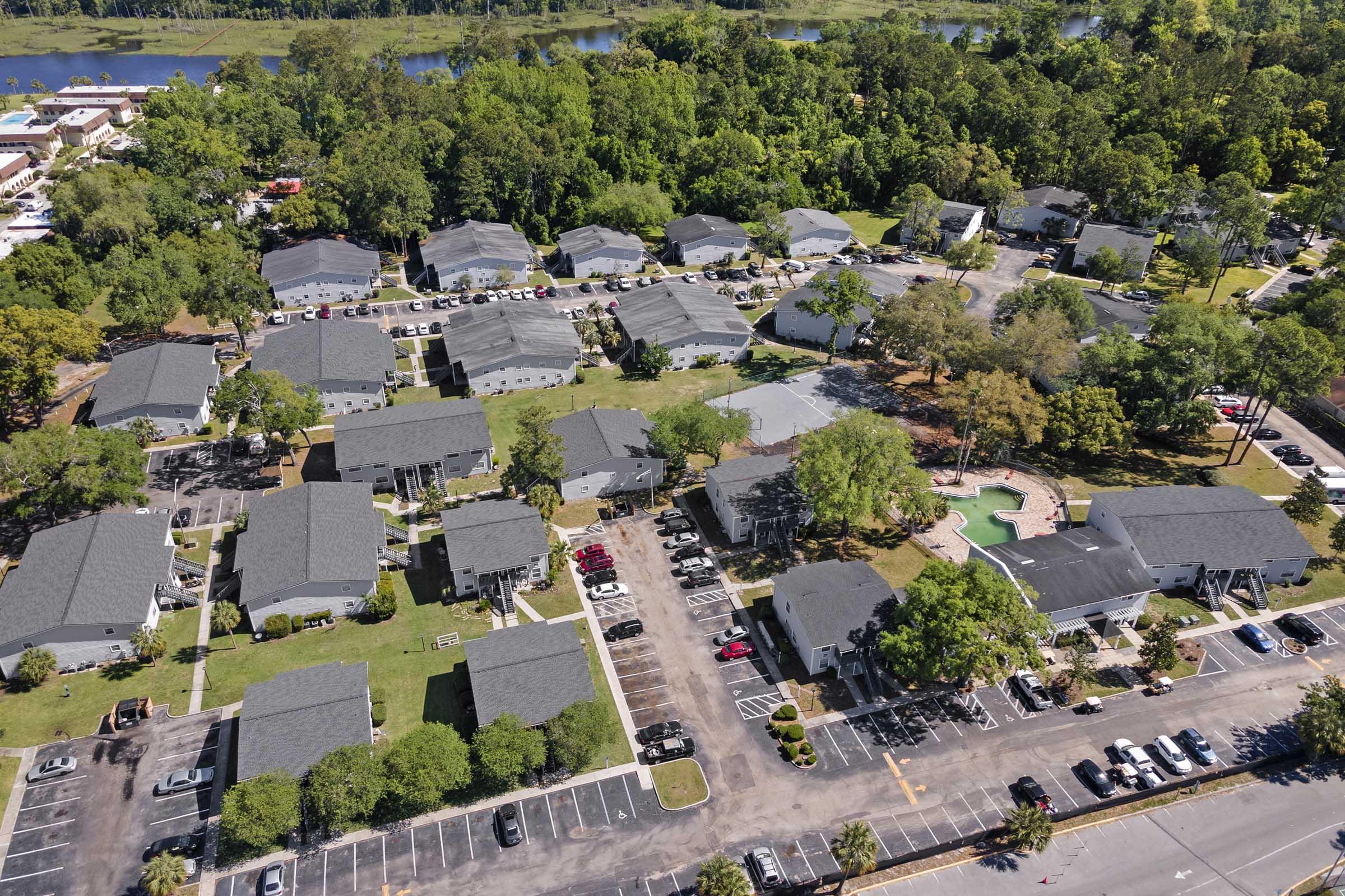 Aerial view of a residential area featuring multiple low-rise buildings arranged in a neighborhood layout. The scene includes a swimming pool surrounded by greenery, parking spaces, and a glimpse of a river in the background, suggesting a peaceful suburban environment.