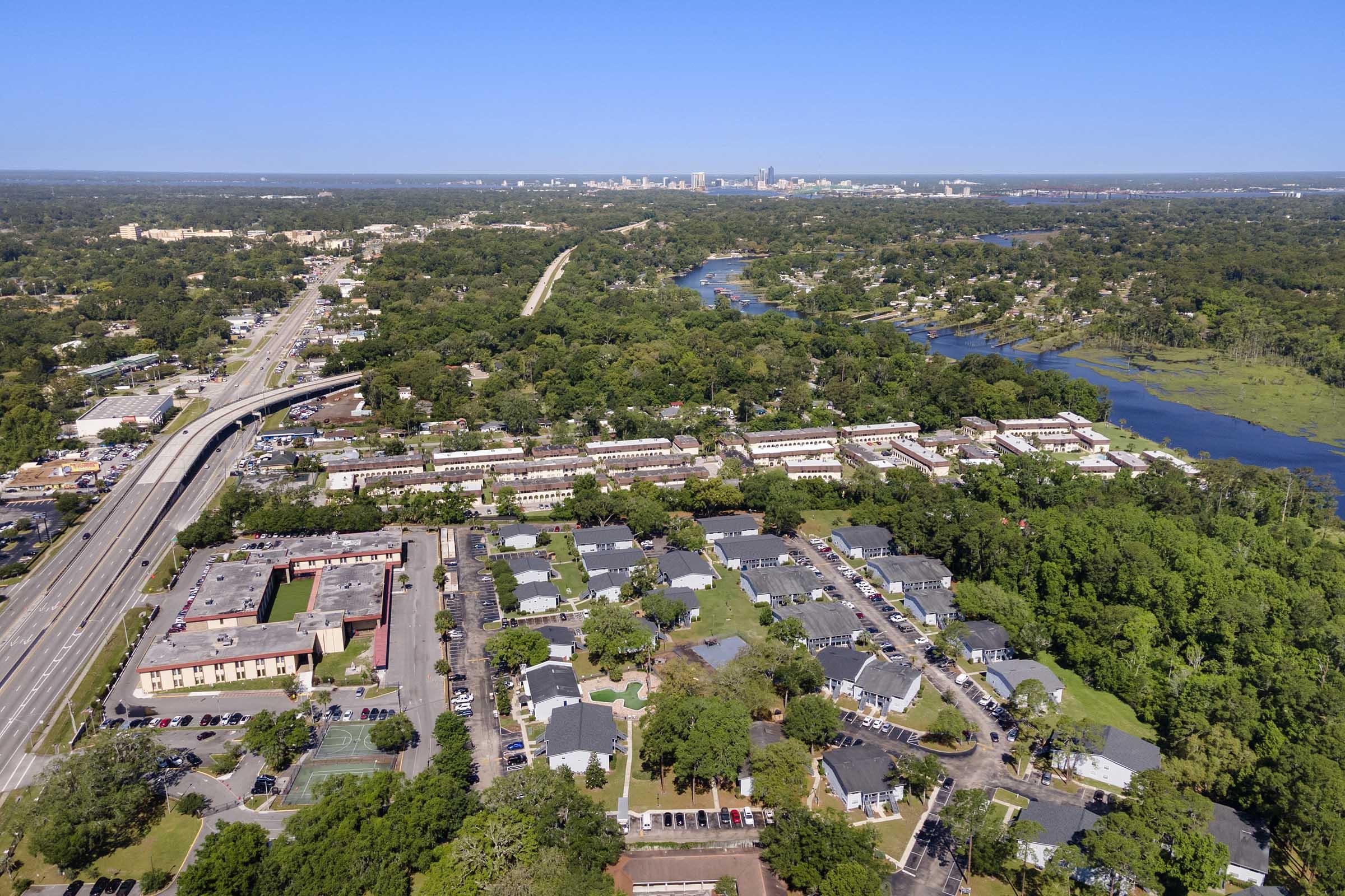Aerial view of a suburban area featuring residential buildings, green trees, and a winding river. In the background, a city skyline is visible along with a highway. The landscape shows a mix of developed and natural spaces, highlighting the contrast between urban and rural environments.