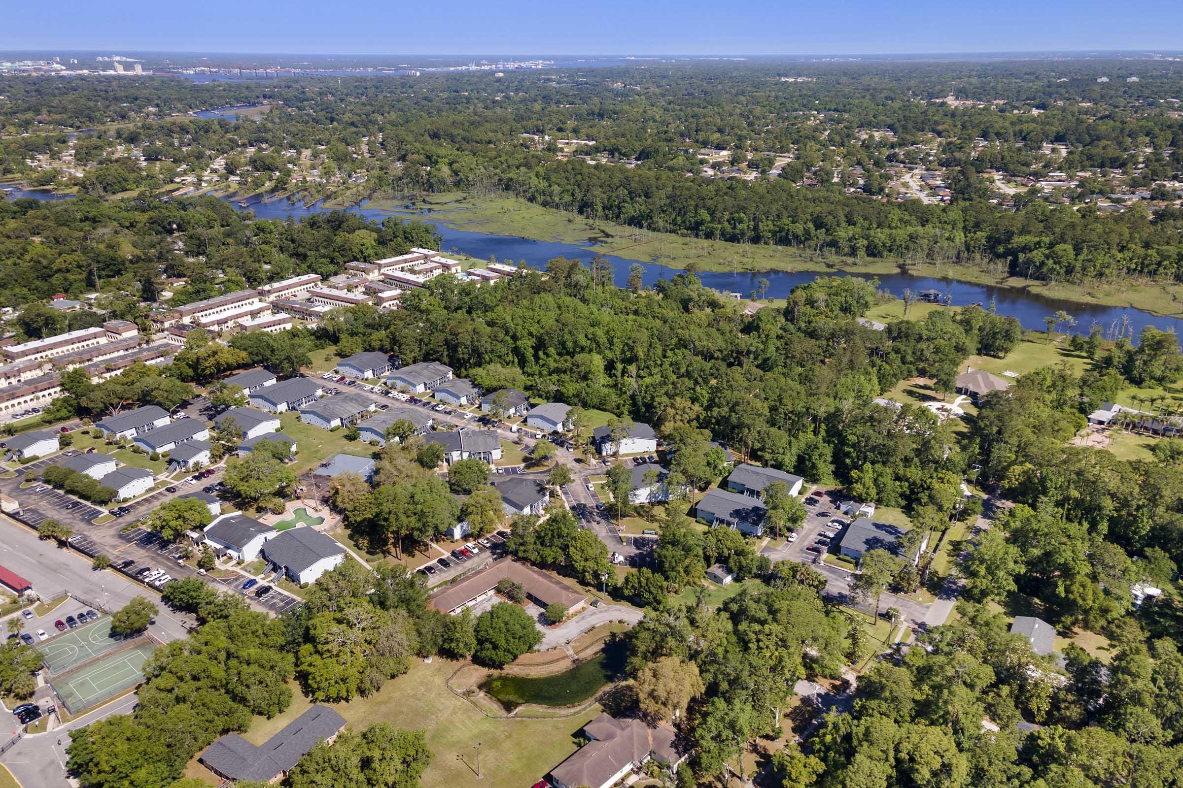 Aerial view of a suburban area featuring residential buildings, lush greenery, and winding waterways. The landscape includes parks, tennis courts, and a mix of developed spaces and natural surroundings, showcasing a harmonious blend of urban and nature in a sunny environment.