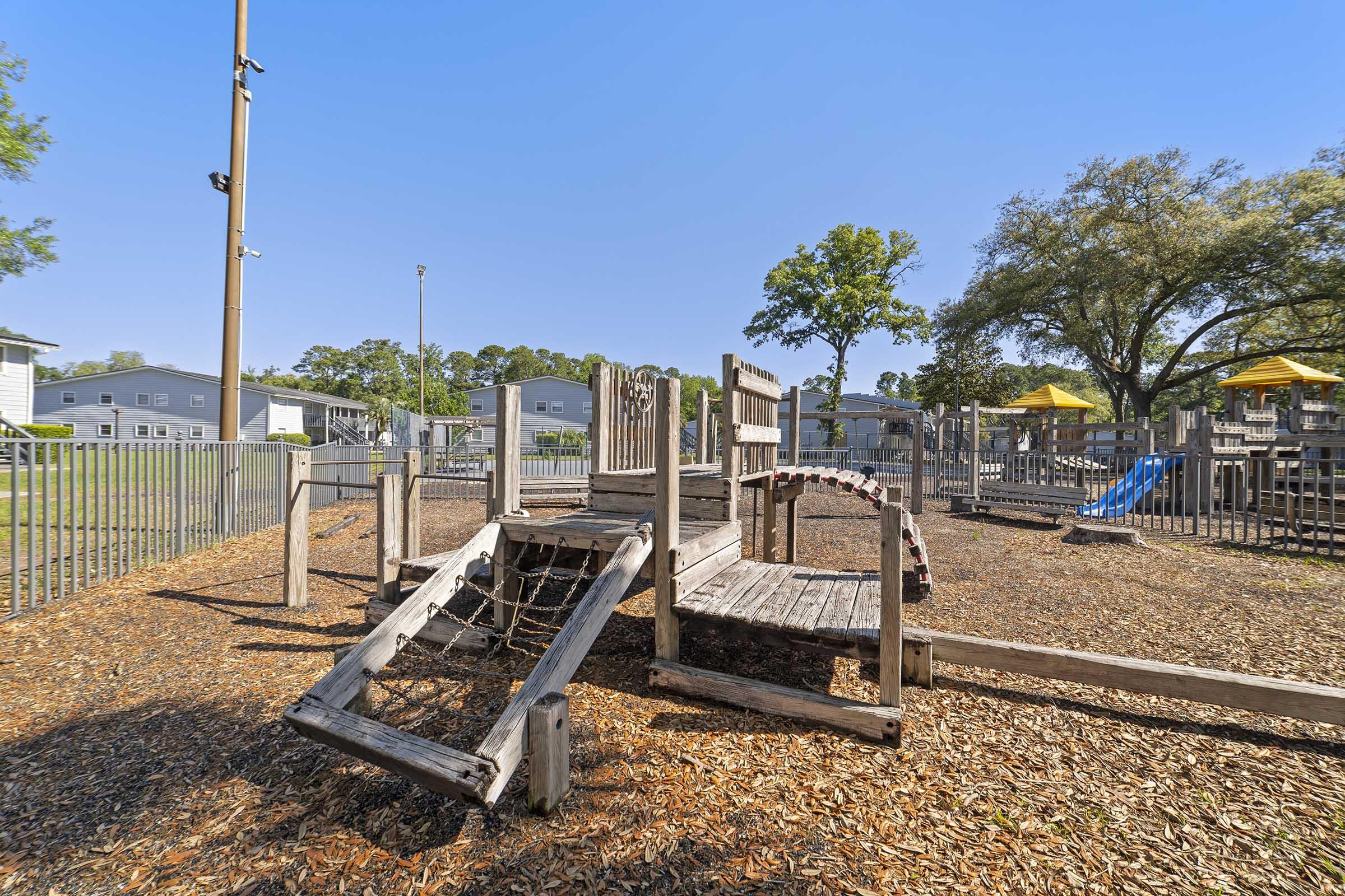 Playground area featuring wooden play structures with climbing elements, a slide, and a rope net. Surrounding the playground is a fenced area with mulch flooring. In the background, there are residential buildings and yellow canopies providing shade. The sky is clear and blue.