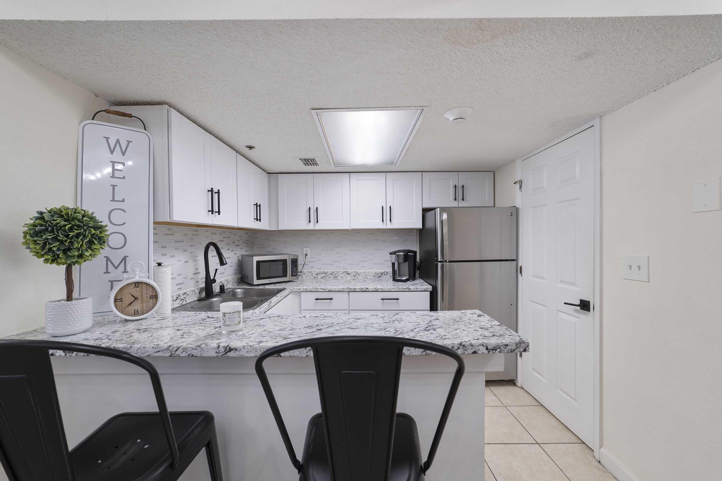 Modern kitchen with white cabinetry and granite countertops. Includes a small potted plant, a clock, and kitchen appliances like a microwave and coffee maker. Two black barstools are positioned at the breakfast bar, and there is a welcoming sign on the wall. Bright lighting enhances the space.