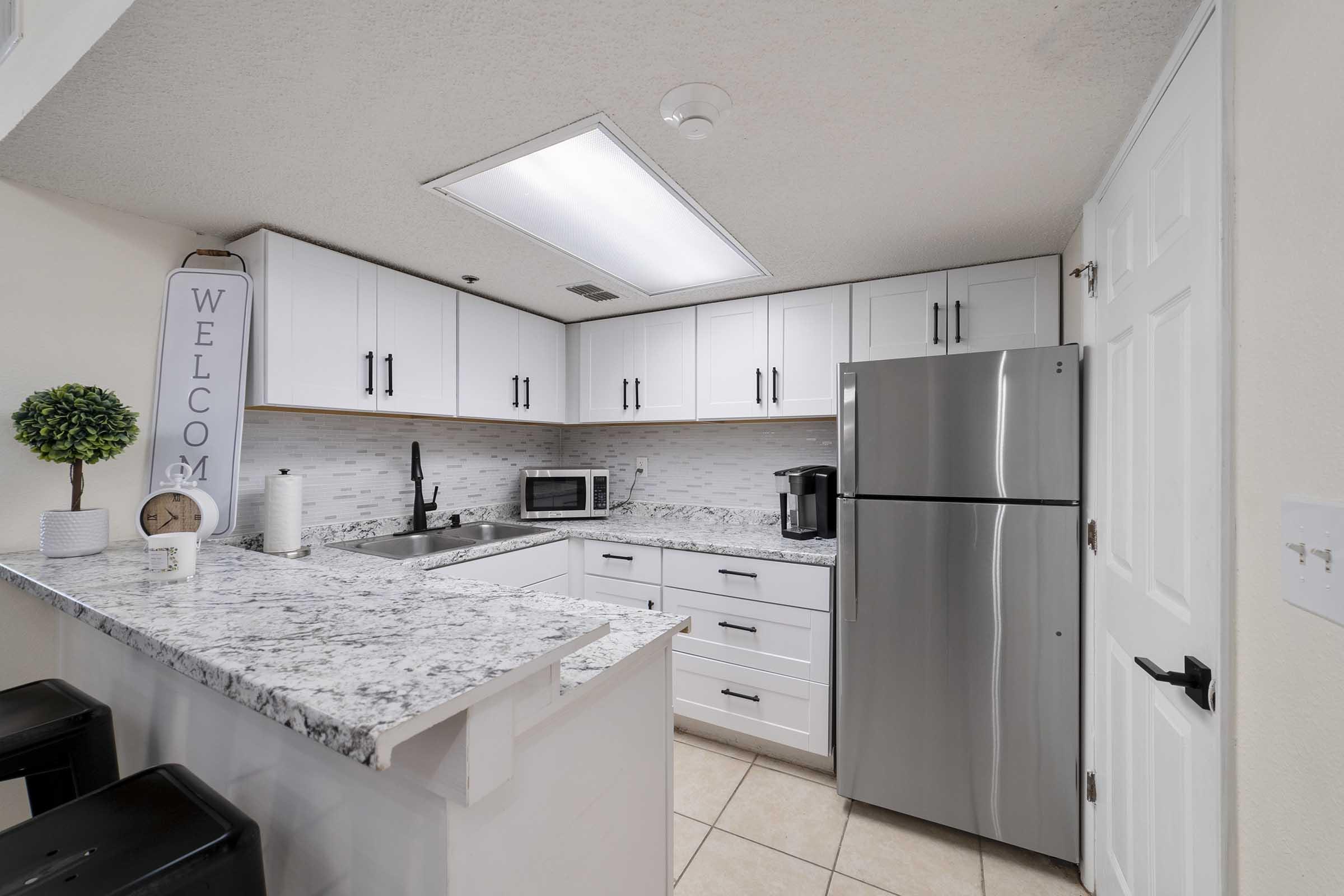Modern kitchen with white cabinetry, a gray granite countertop, and stainless steel appliances, including a refrigerator and microwave. The space features a decorative welcome sign and a small potted plant on the countertop, with neutral tile flooring and a bright overhead light.