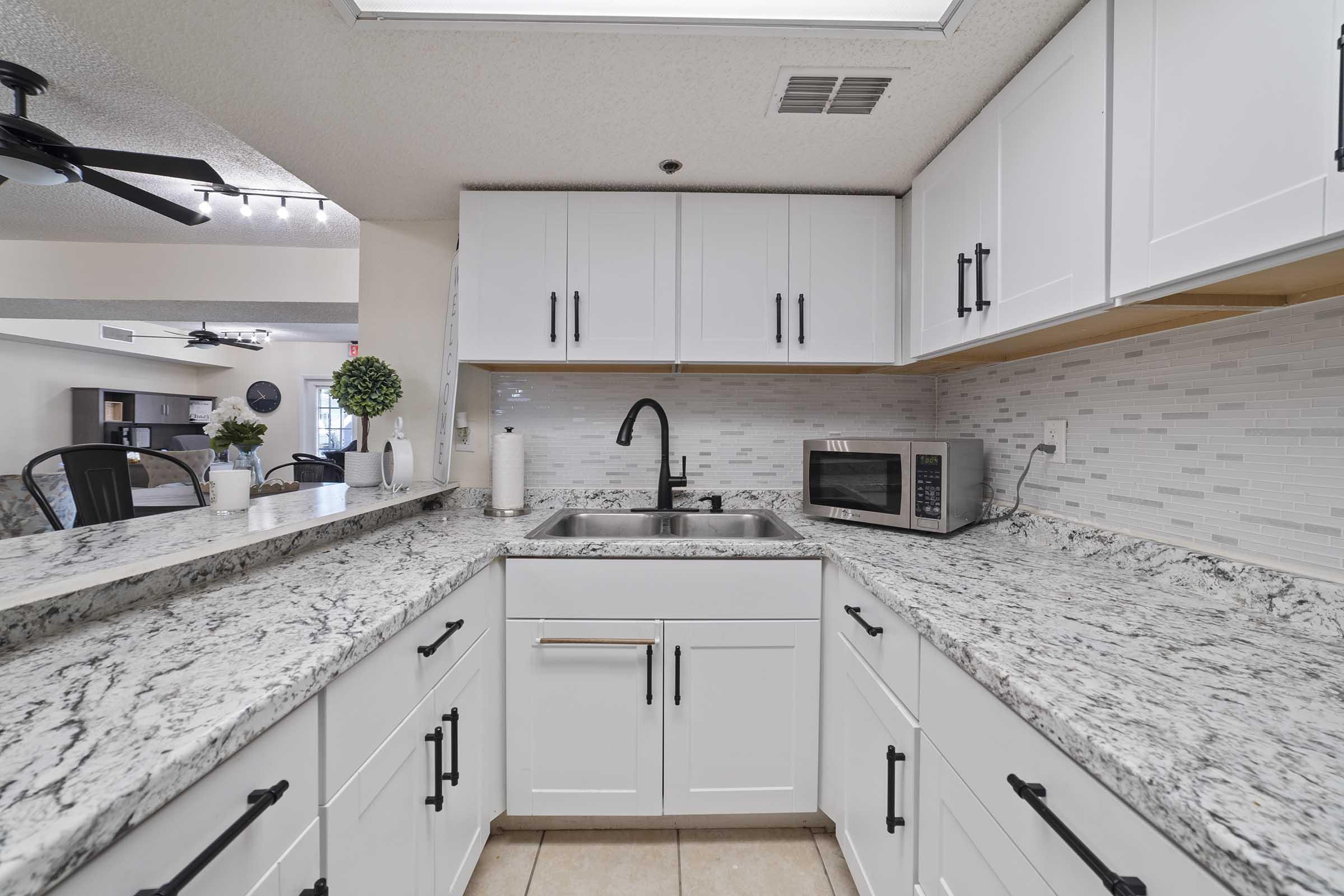 A modern kitchen featuring white cabinets with black handles, a granite countertop, a stainless steel sink, and a microwave. The backsplash consists of light-colored tiles. A potted plant adds a touch of greenery, and the space is well-lit, creating a clean and inviting atmosphere.