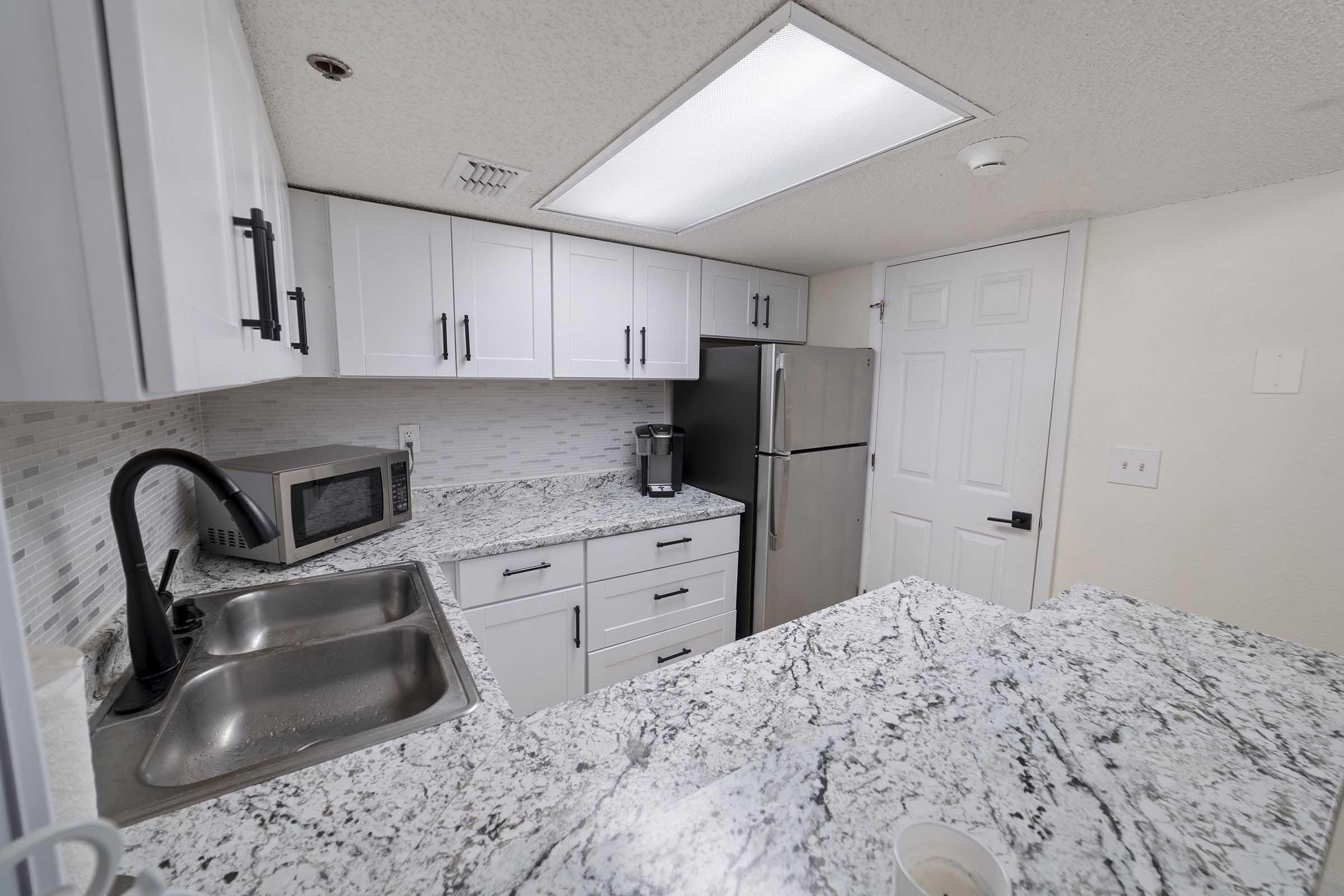 Modern kitchen with white cabinetry, granite countertops, and stainless steel appliances. Features include a dual-basin sink, a microwave, and a refrigerator. The backsplash consists of light tiles, and the room is well-lit with ceiling lighting. A door leads to another area.