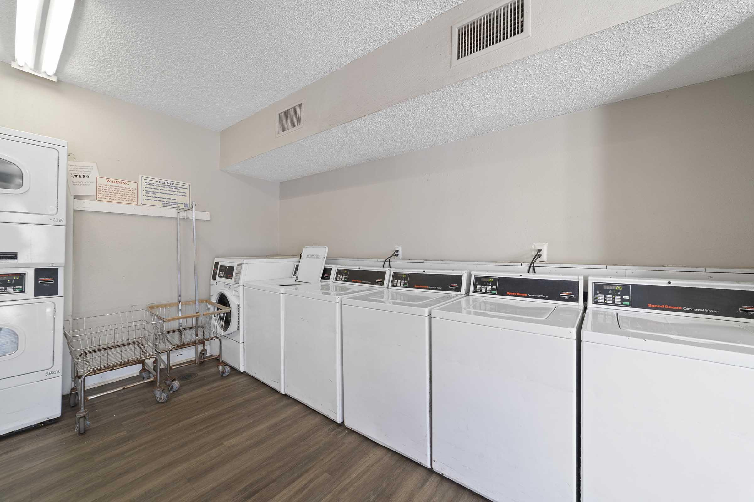 A laundry room featuring several white washing machines and dryers lined up against a wall. There is a folding table and a laundry cart nearby. The space has gray walls and a wooden floor, illuminated by fluorescent lights on the ceiling.