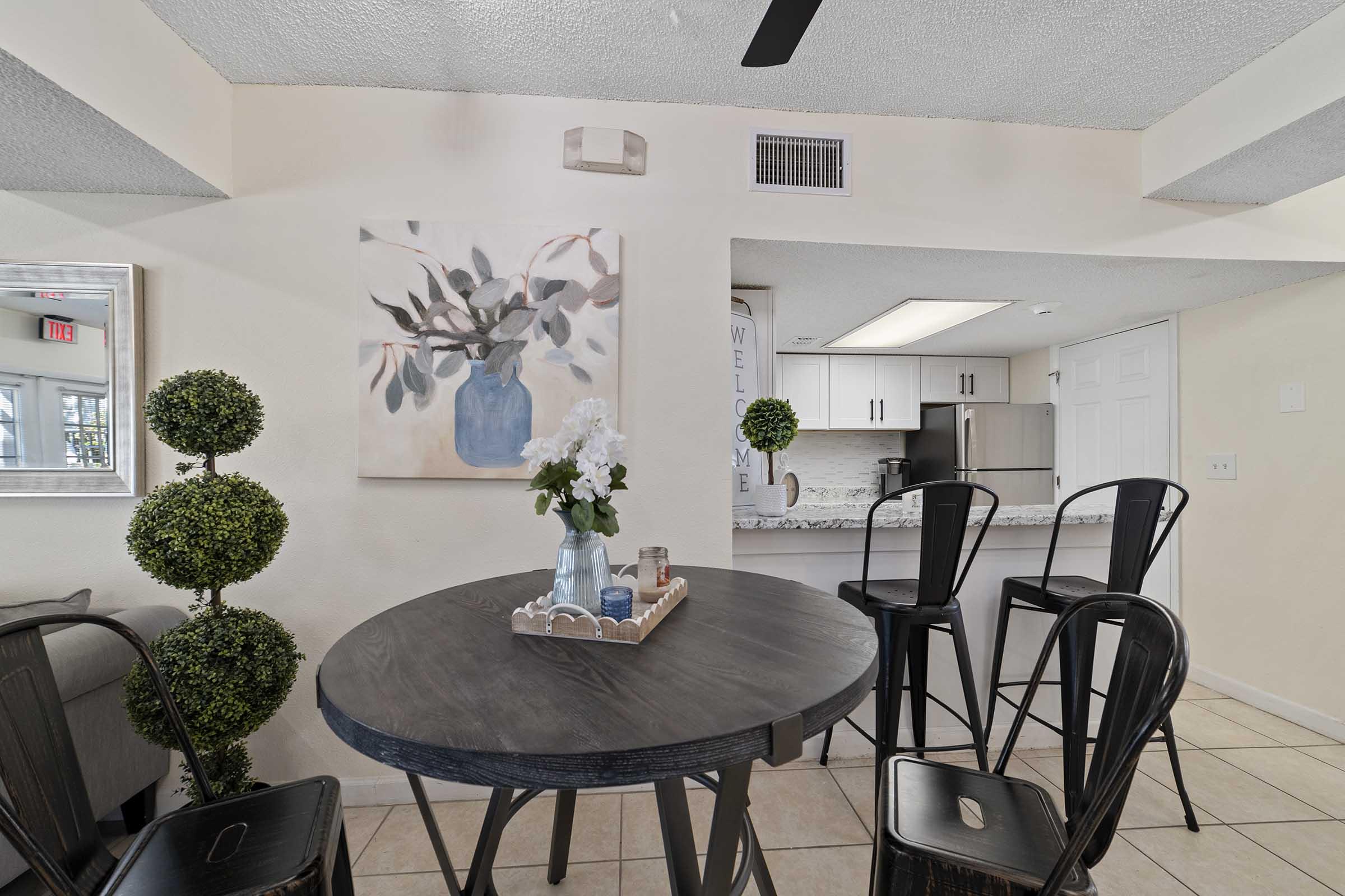 A cozy dining area featuring a round black table surrounded by black chairs. A decorative arrangement includes a small vase with white flowers and two decorative topiaries. The background shows a modern kitchen with light-colored cabinets and a welcome sign on the wall, creating a warm and inviting atmosphere.