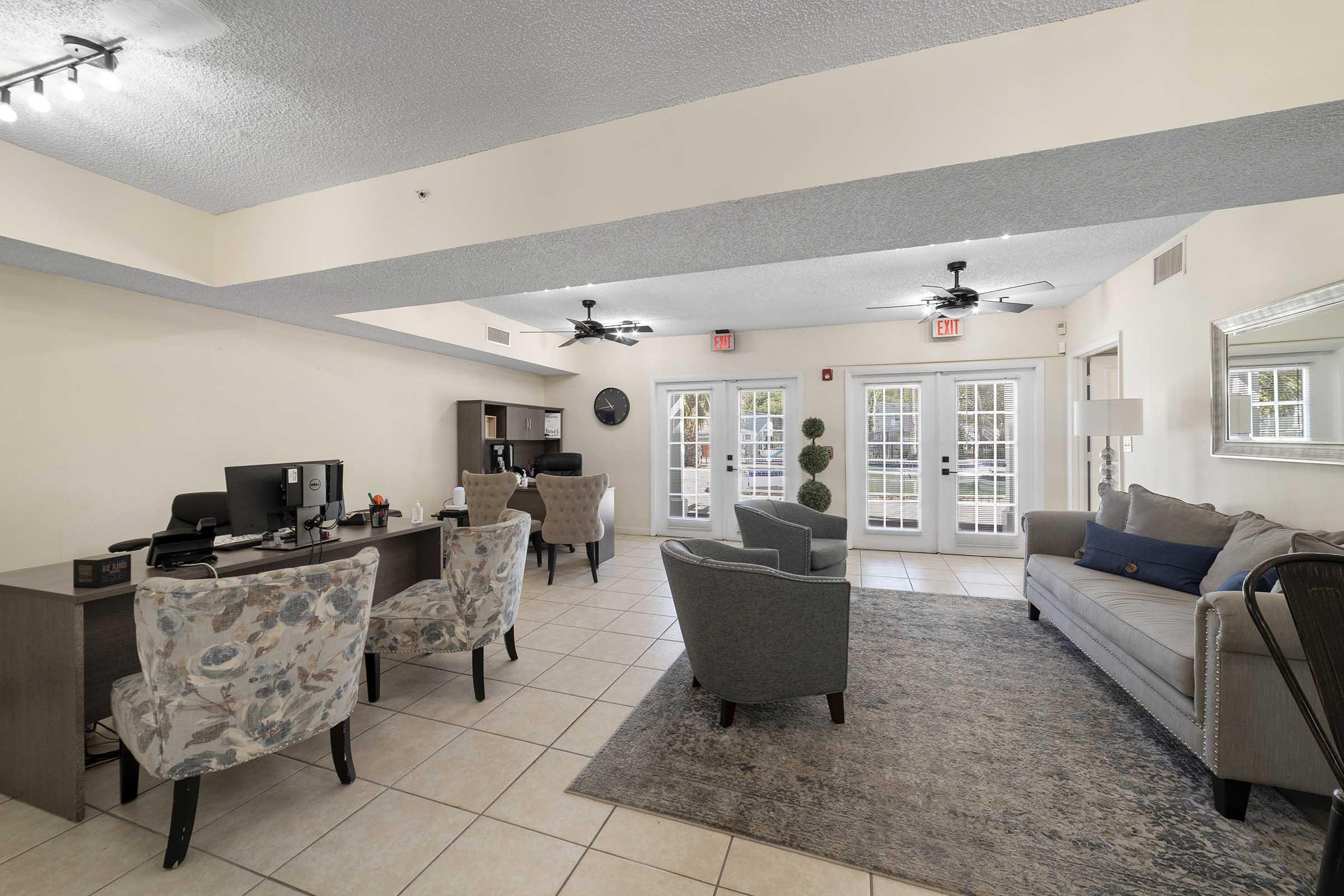 Interior view of a modern lounge area featuring two patterned chairs and a grey sofa. There are two desks with computers, ceiling fans, and large sliding glass doors leading to an outdoor space. Light-colored walls and a decorative rug complete the welcoming atmosphere.