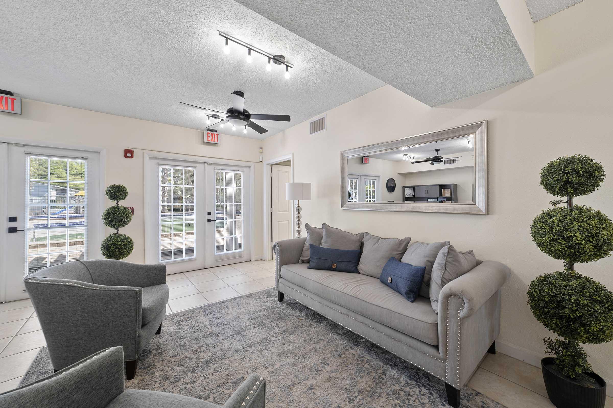 A cozy living room area featuring a light gray sofa with decorative pillows, two armchairs, and a large mirror. Natural light streams in through double glass doors that lead outside, where greenery is visible. Decorative topiary plants add a touch of elegance to the space. A ceiling fan is installed for comfort.