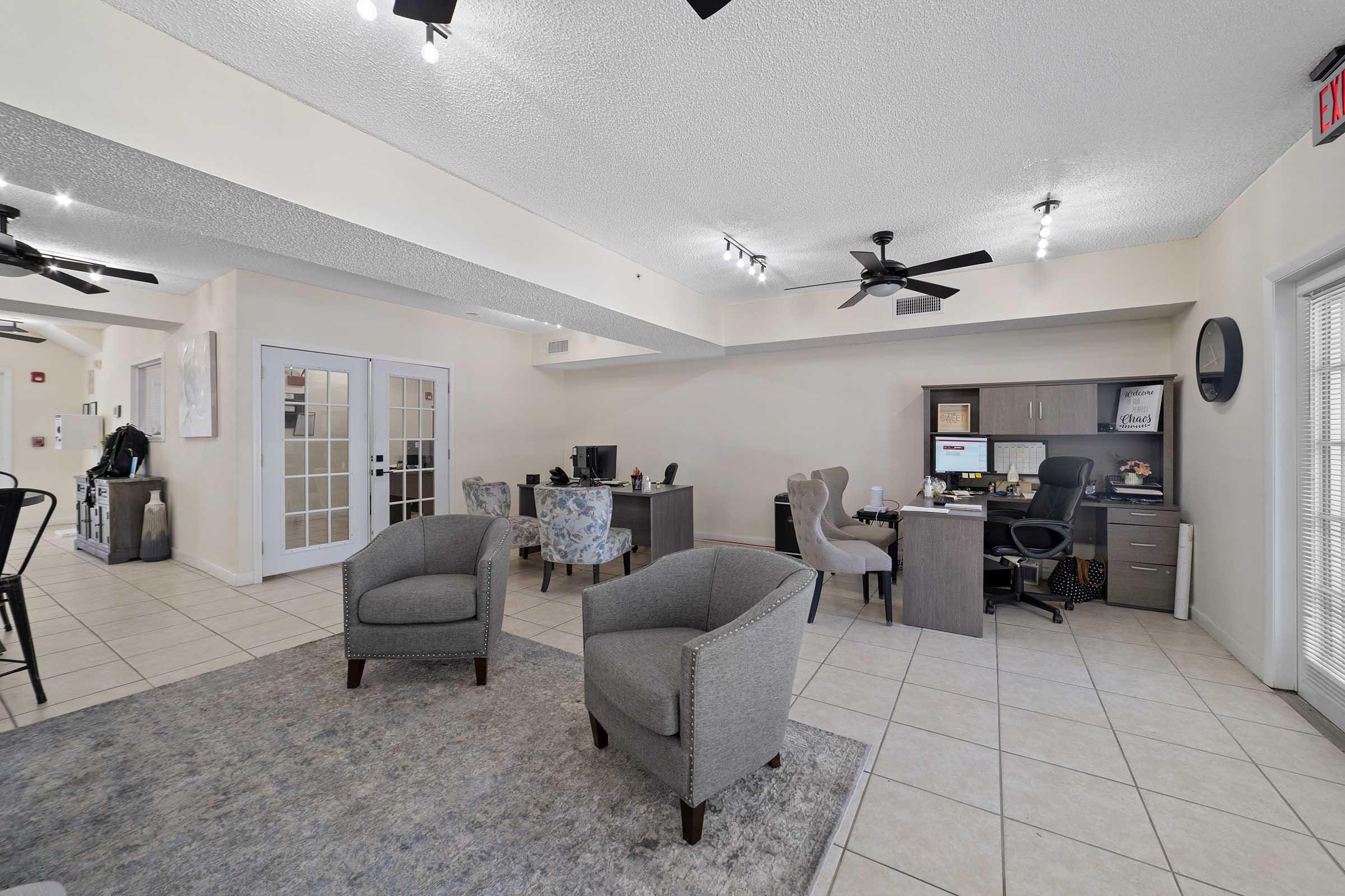 A modern office space featuring two gray armchairs, a desk with a computer, and a chair in a neutral-toned room. The area has tiled flooring, subtle lighting from ceiling fixtures, and a rug in the center, creating a professional yet inviting atmosphere.