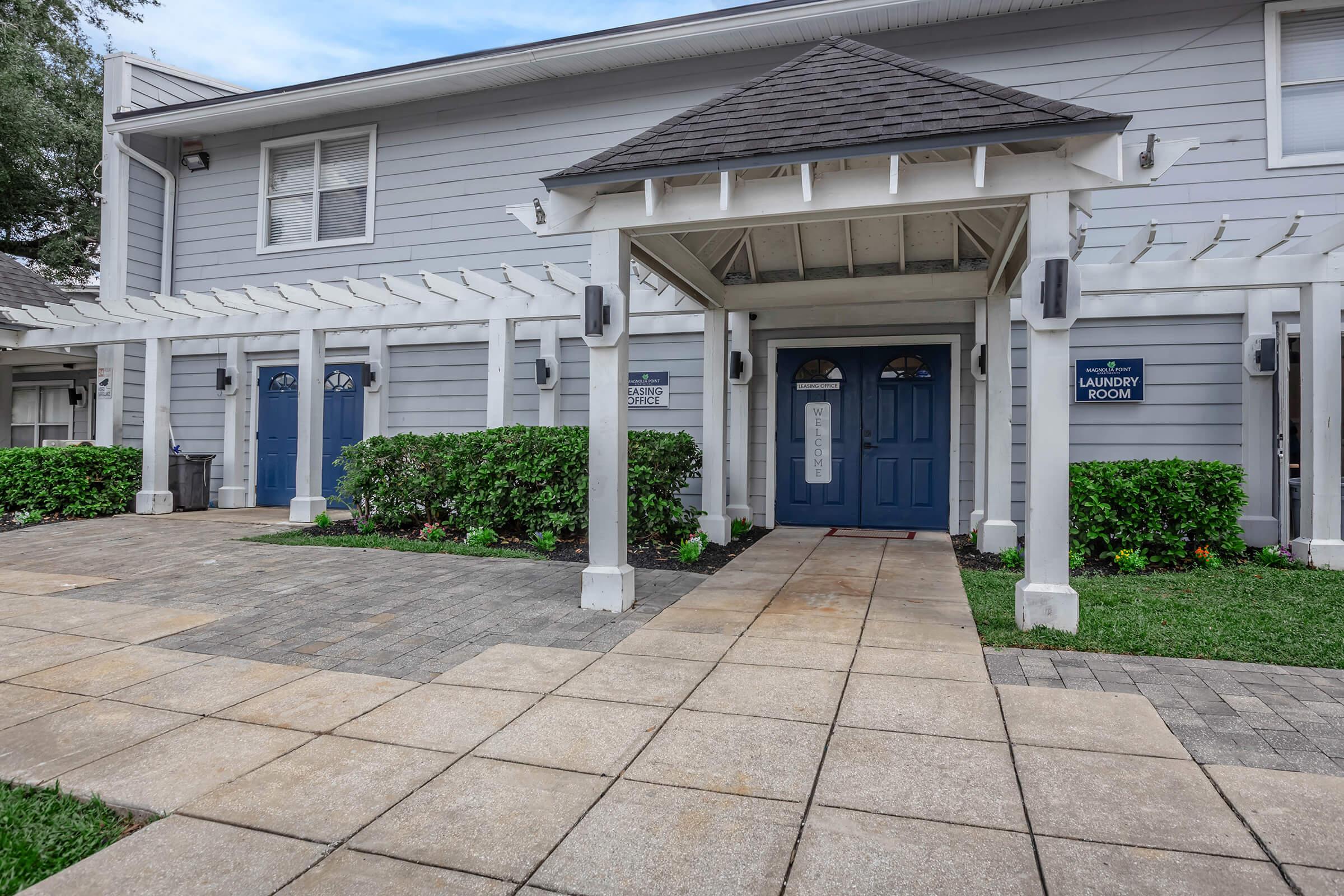 Exterior view of a building featuring a covered entrance with two blue doors labeled "Office" and "Laundry Room." The pathway is made of square tiles, and there is neat landscaping with greenery on both sides. The building has a light gray exterior with a gabled roof, enhancing its welcoming appearance.