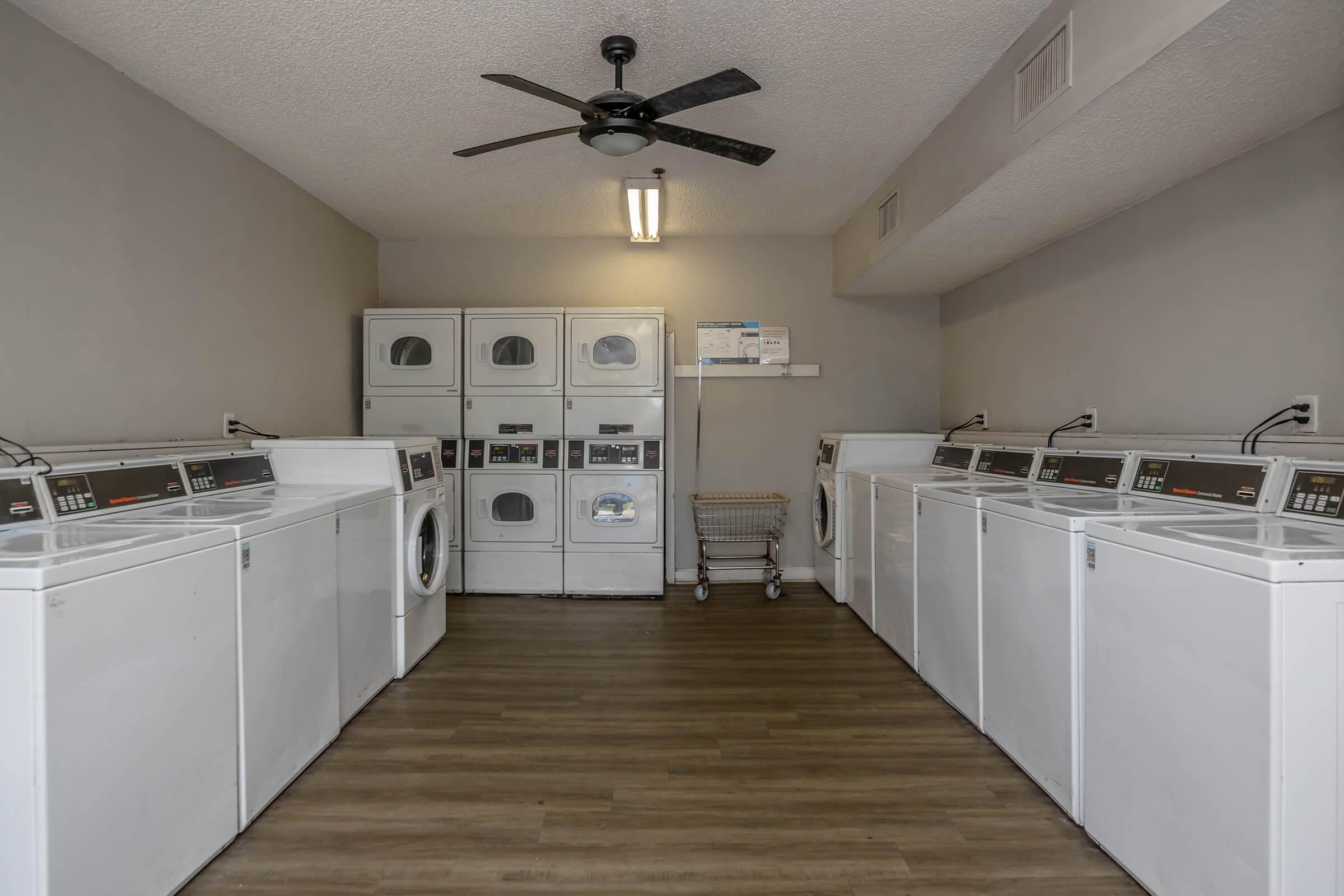 A spacious laundry room with multiple white washing machines and dryers arranged along the walls. There's a large folding table in the center and a ceiling fan above. The floor is hardwood, and the walls are painted a light gray, creating a clean and modern atmosphere. A cart for laundry is also visible.