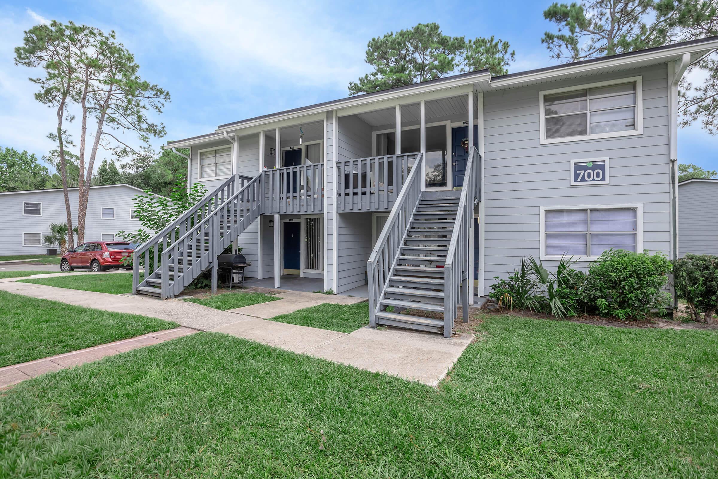 Two-story apartment building featuring a gray exterior, stairs leading to the second floor, and a blue door on the upper level. The front yard is grassy with a sidewalk, and there are nearby trees. A red car is parked in the lot to the left, and additional apartment buildings can be seen in the background.