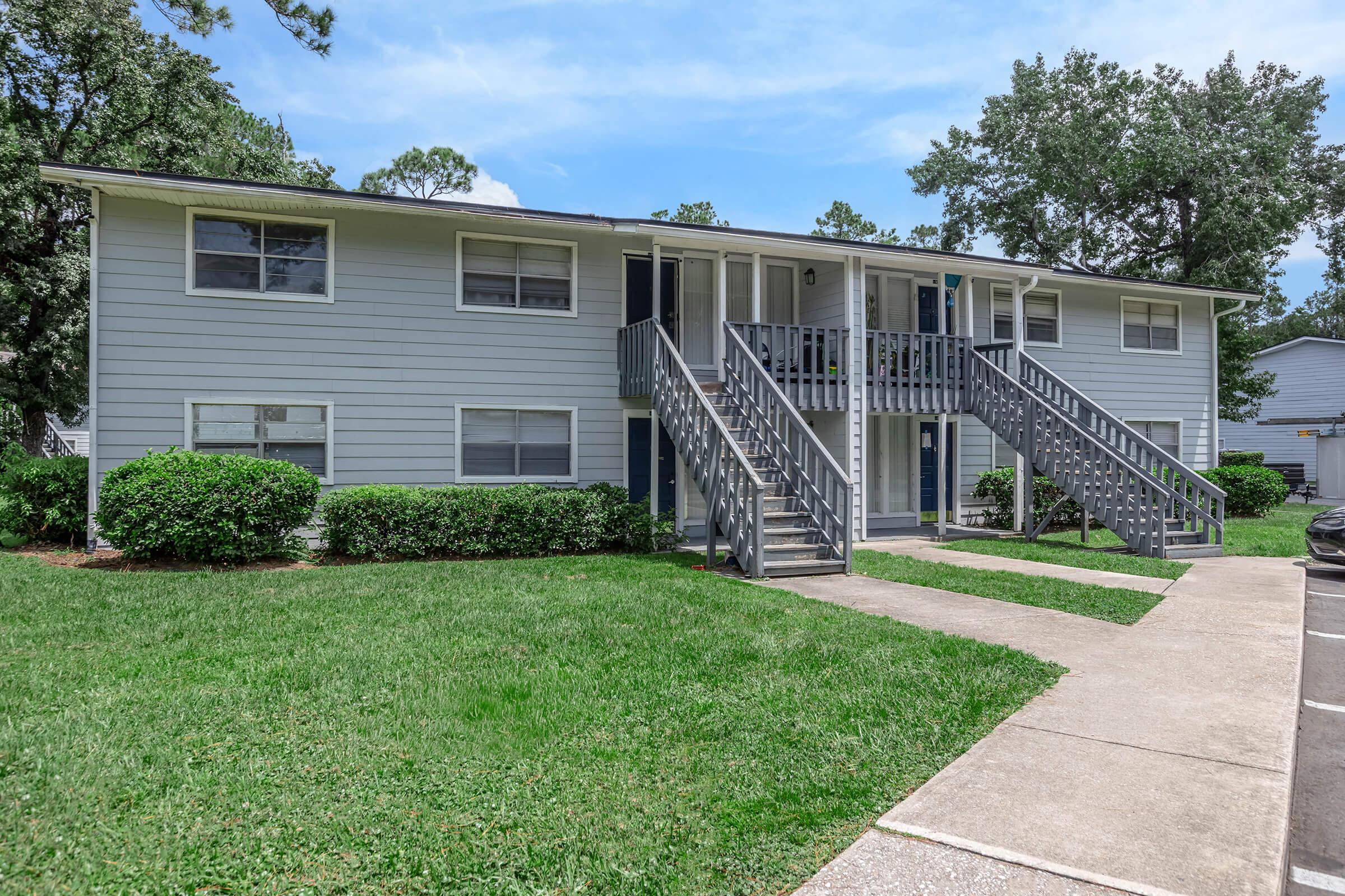 A two-story residential building with gray siding, featuring multiple windows and two sets of outdoor stairs. The building is surrounded by well-maintained grass and shrubs. There are parking spaces visible in front of the building. The sky is clear with a few clouds.