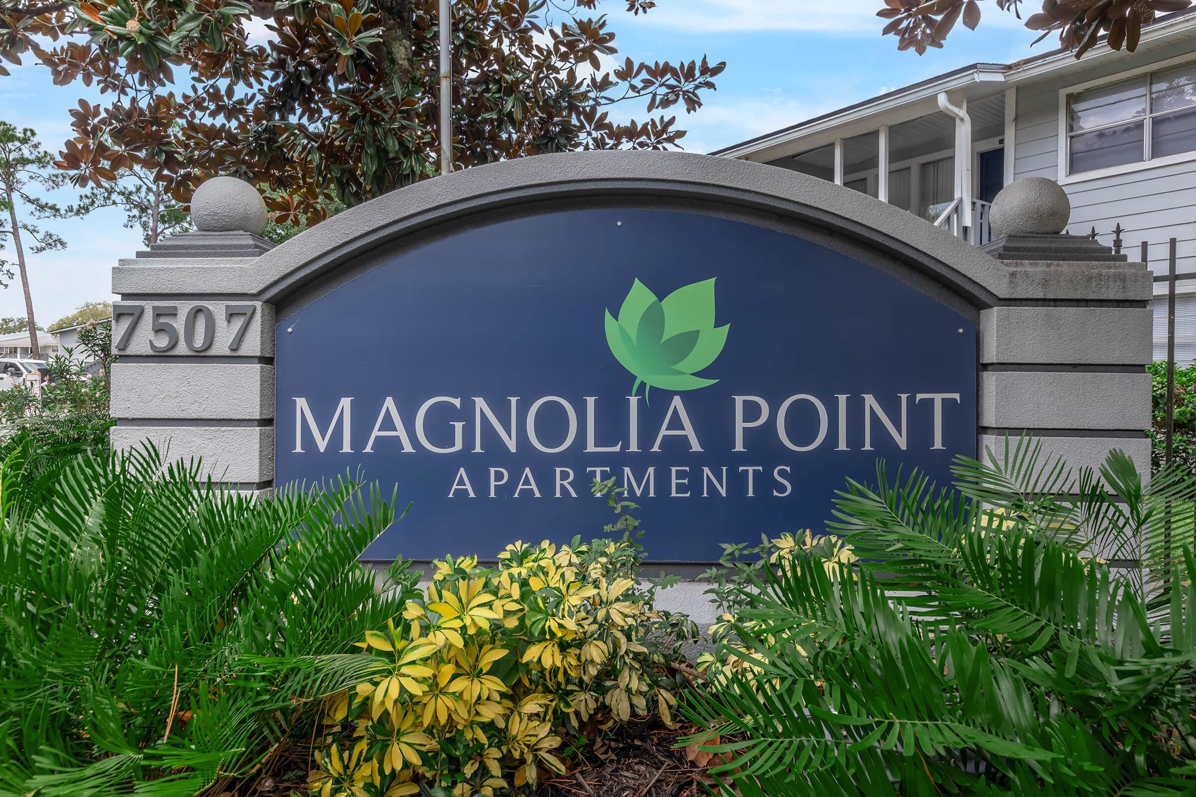 Sign for Magnolia Point Apartments, featuring a green lotus logo above the name. The sign is framed by lush green plants and shrubs, indicating a welcoming entrance. The address "7507" is displayed prominently at the bottom.