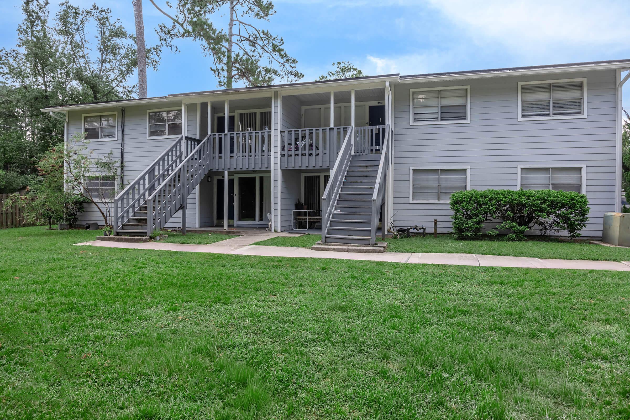 Two-story residential building with a gray exterior and white trim, featuring a grassy front yard. Stairs lead to the upper level from both sides of the porch. There are shrubs and trees in the background, and a concrete pathway in front of the building. Clear blue sky above.
