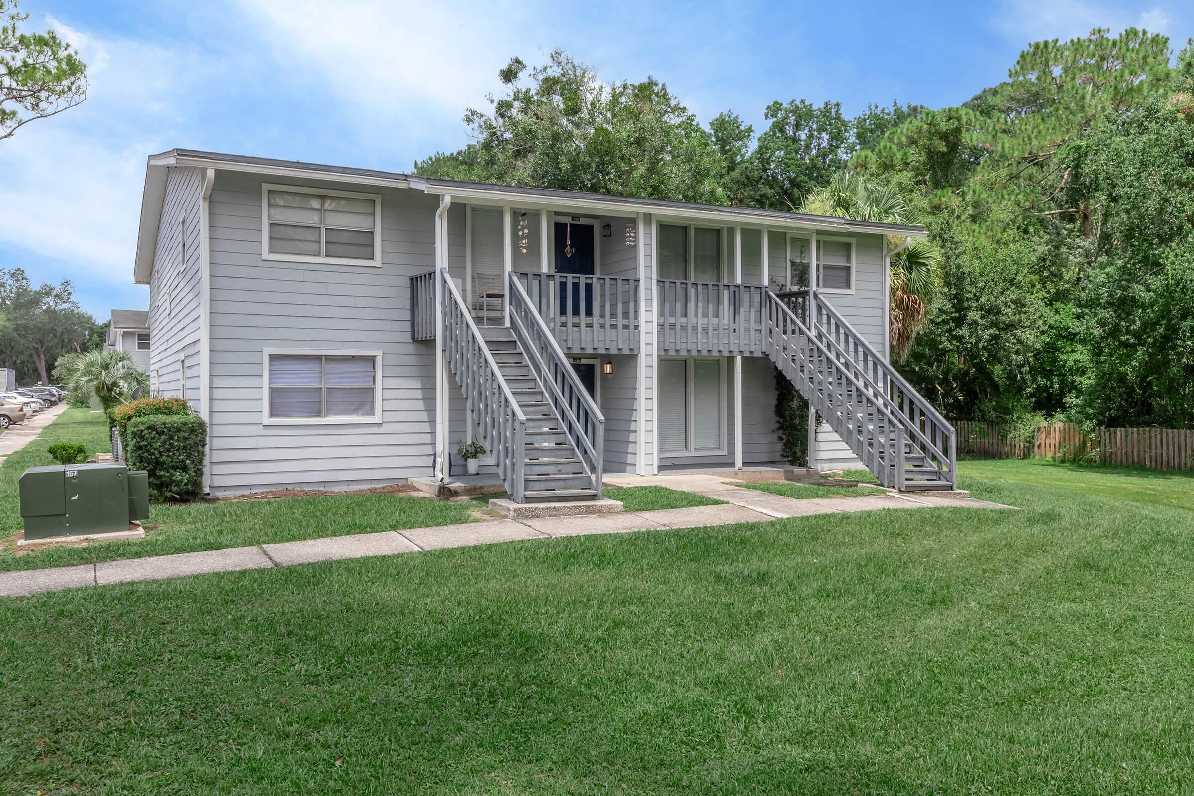 A two-story apartment building with a light gray exterior and two sets of stairs leading to the second floor. The building is surrounded by green grass and trees, with a pathway in front. There is a utility box on the side and a wooden fence in the background. The sky is clear and blue.