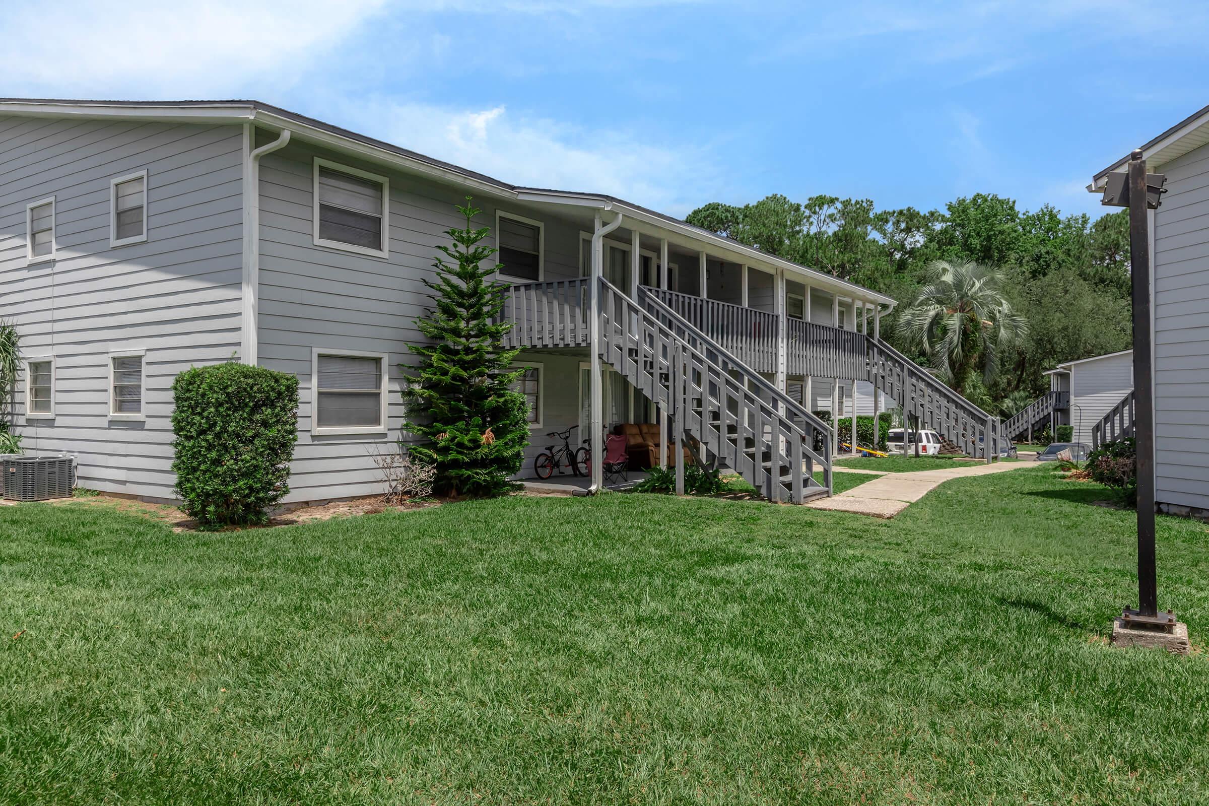 A two-story apartment building with gray siding, featuring multiple staircases leading to the upper level. The surrounding area includes well-maintained green grass, small shrubs, and trees in the background. A bike is parked near the entrance, giving a welcoming residential vibe.
