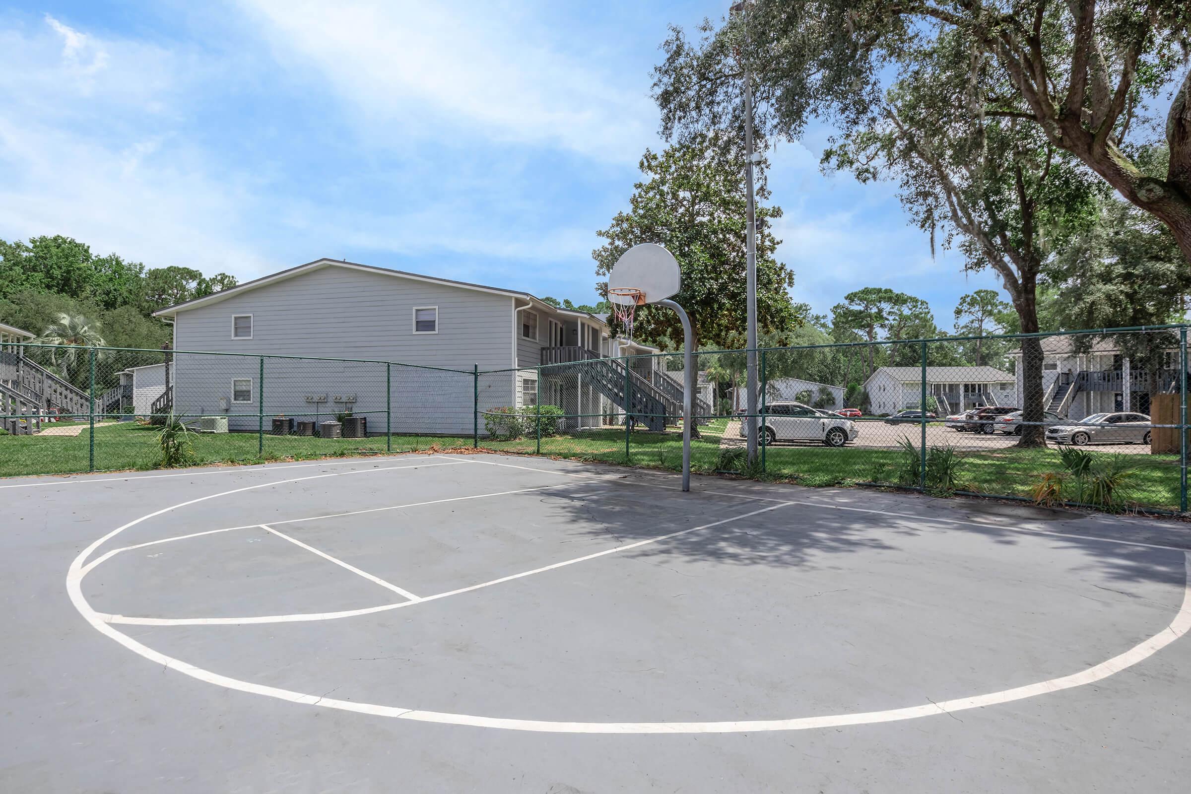 A well-maintained outdoor basketball court with a clear sky in the background. The court is enclosed by a fence and features a basketball hoop. In the background, there are residential buildings surrounded by trees and a parking area with cars. The scene suggests a recreational space in a community setting.