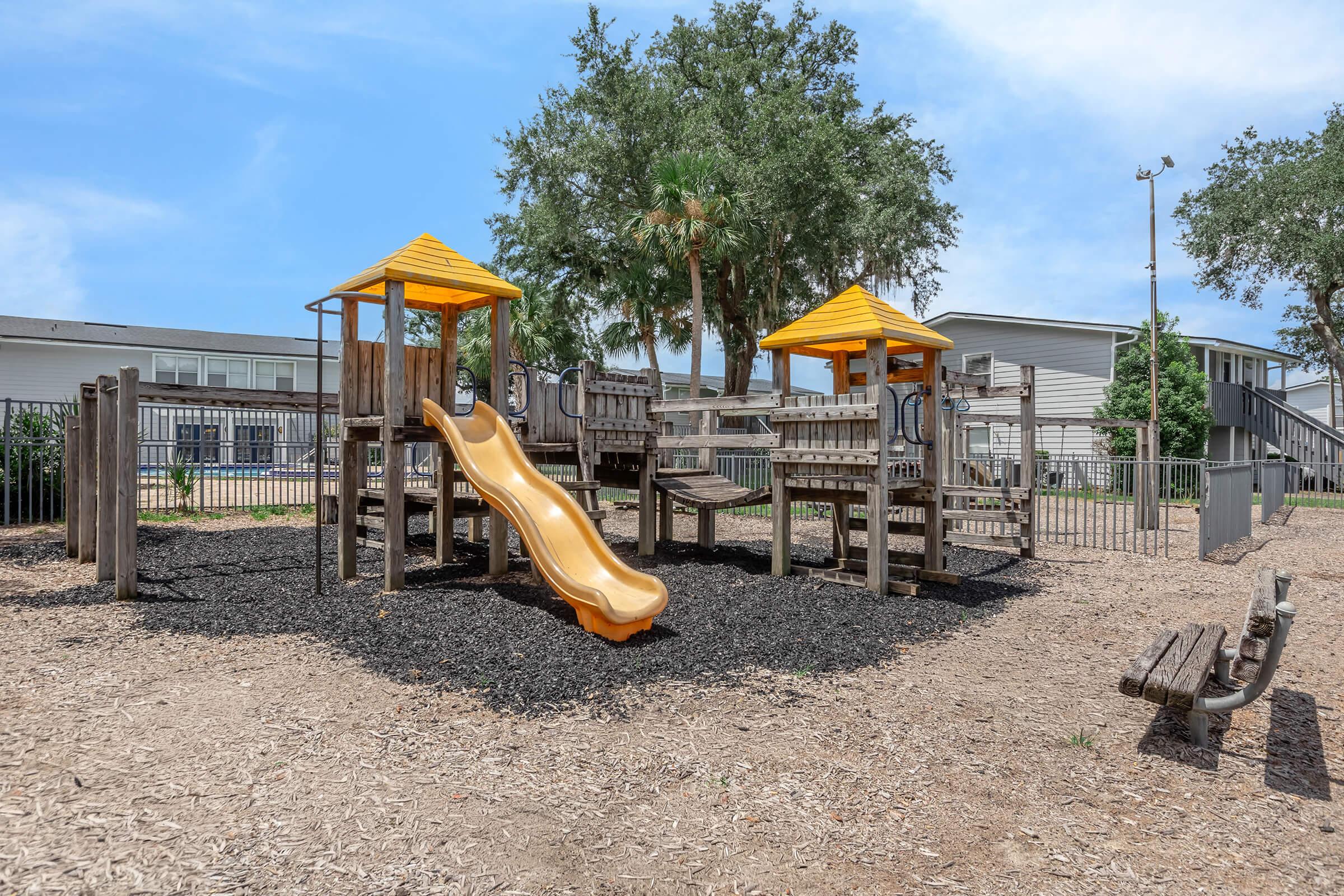 A playground featuring two yellow-roofed playhouses, a yellow slide, and various climbing structures surrounded by black mulch. Benches are visible nearby, and residential buildings are in the background under a clear blue sky.