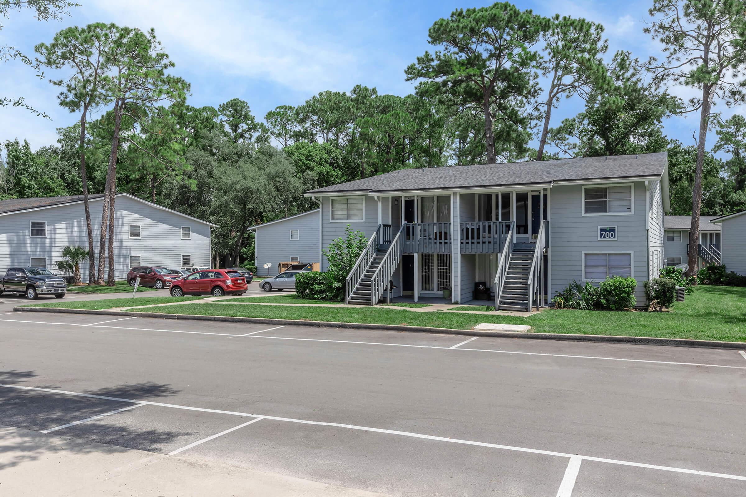 Two-story residential building with multiple apartments, featuring a grey exterior and large stairs leading to the upper level. Green landscaping surrounds the building, with several parked cars visible in the parking lot. The setting is serene, with trees in the background and a clear blue sky.