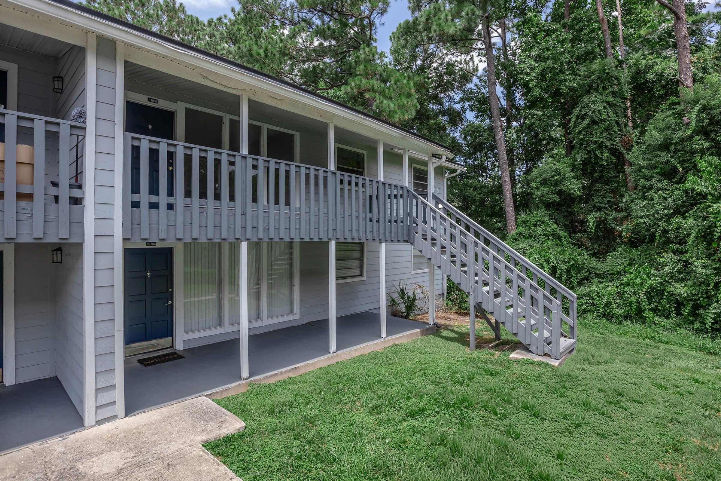 A two-story building with a gray exterior featuring a set of stairs leading to the upper level. The ground floor has a blue door and large windows, while the surrounding area includes green grass and trees in the background, creating a serene outdoor setting.