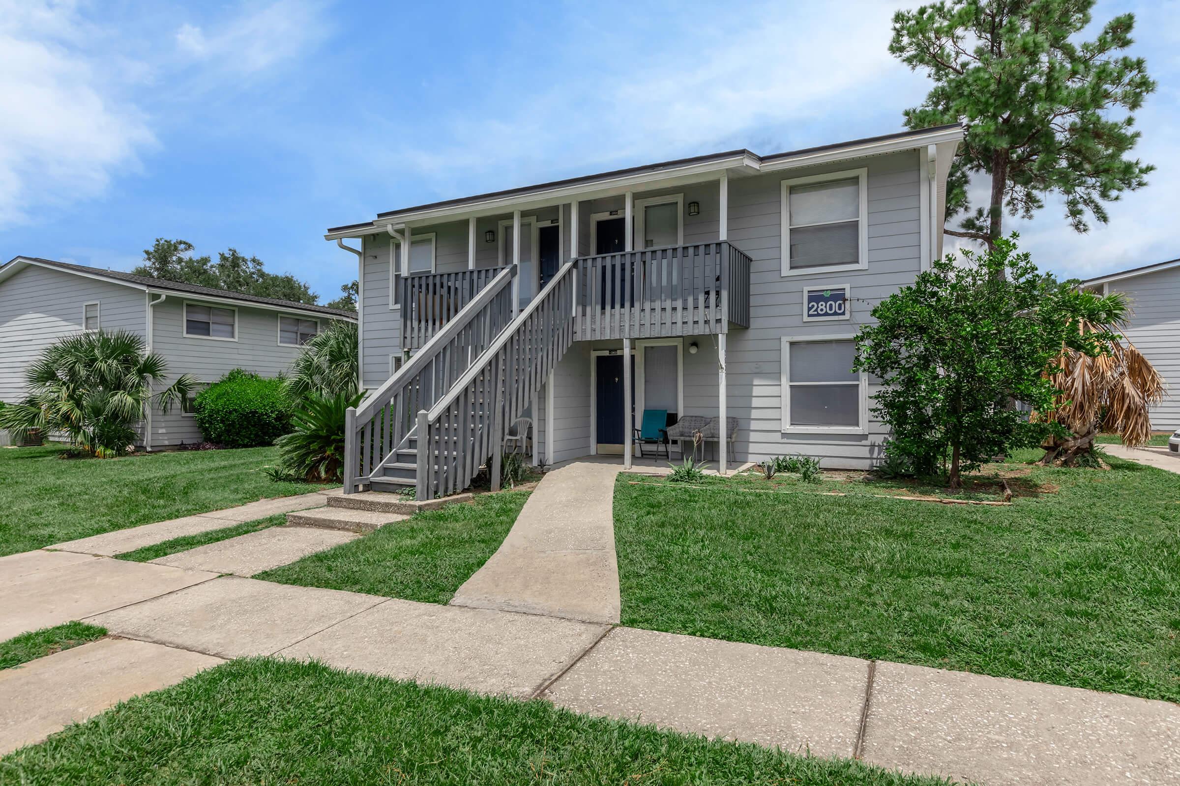 Two-story residential building with gray exterior and white trim. A staircase leads to the second floor, where a small balcony is visible. The surrounding area features well-kept lawns, palm trees, and sidewalks. The building number "2800" is displayed on the front. Clear blue sky overhead.
