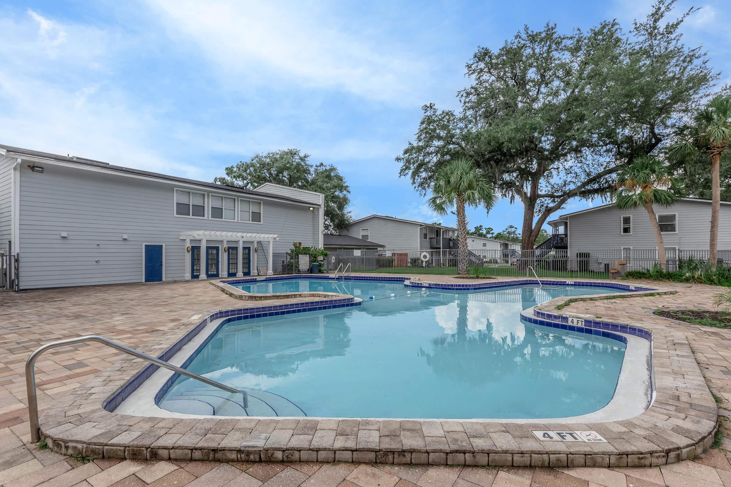 A serene outdoor pool area with a wavy-shaped swimming pool surrounded by pavers. There are lounge chairs and palm trees nearby, with a few buildings in the background. The sky is clear, creating a relaxing atmosphere for leisure and recreation.