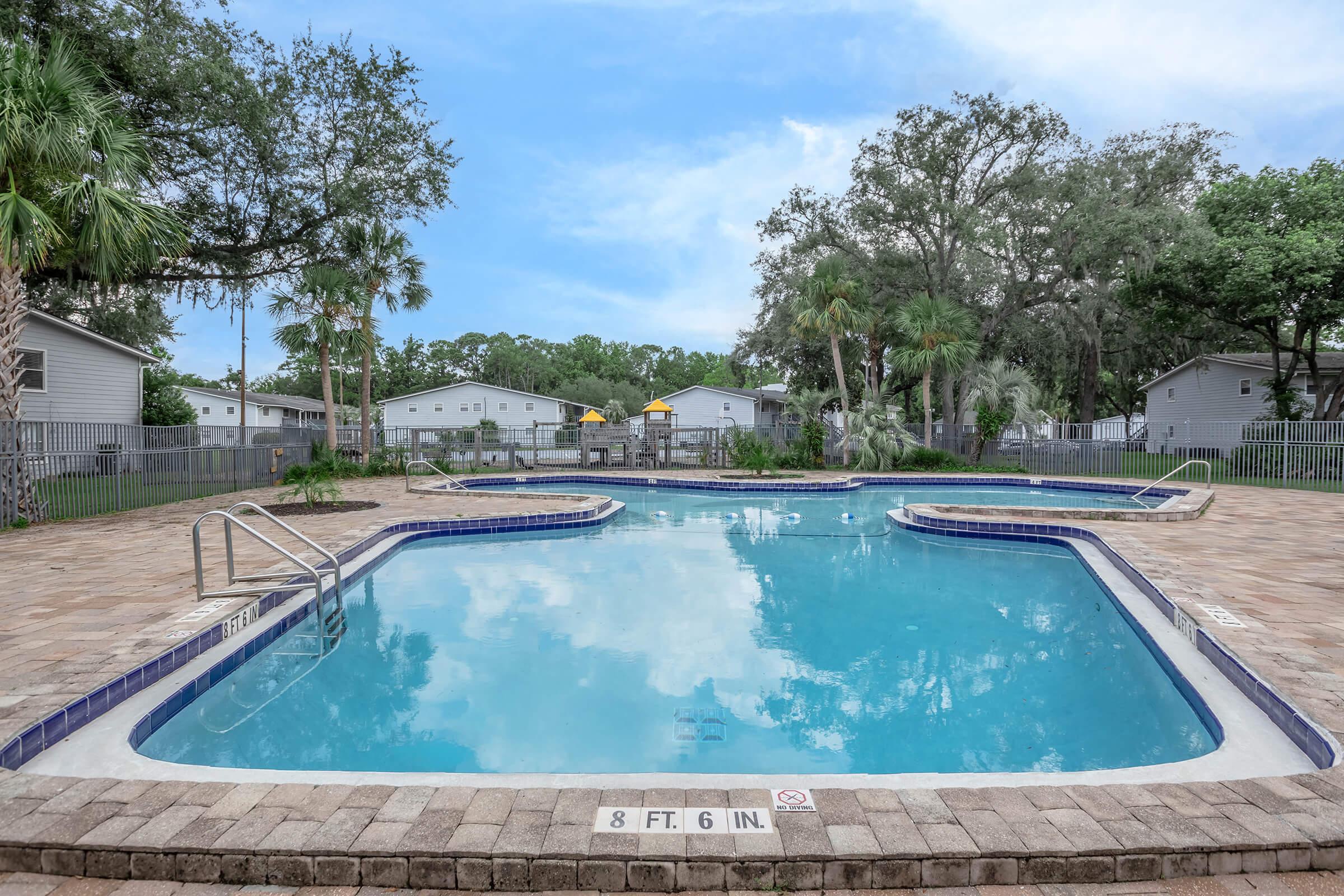 Swimming pool surrounded by palm trees and paved area, with a depth marker indicating 8 feet 6 inches. There are lounge chairs and a fence in the background, with residential buildings visible. The sky is clear with a few clouds, creating a serene outdoor atmosphere.