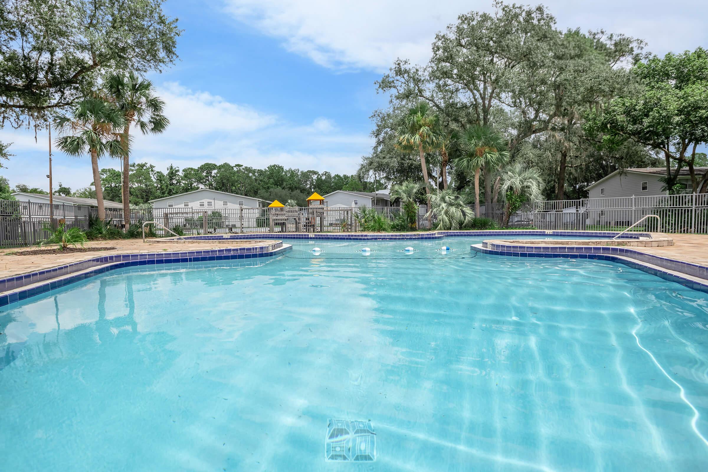 A serene swimming pool surrounded by lush greenery and palm trees, with clear blue water reflecting the sky. In the background, there are residential buildings and playground equipment. The scene conveys a calm and inviting atmosphere for relaxation and leisure.