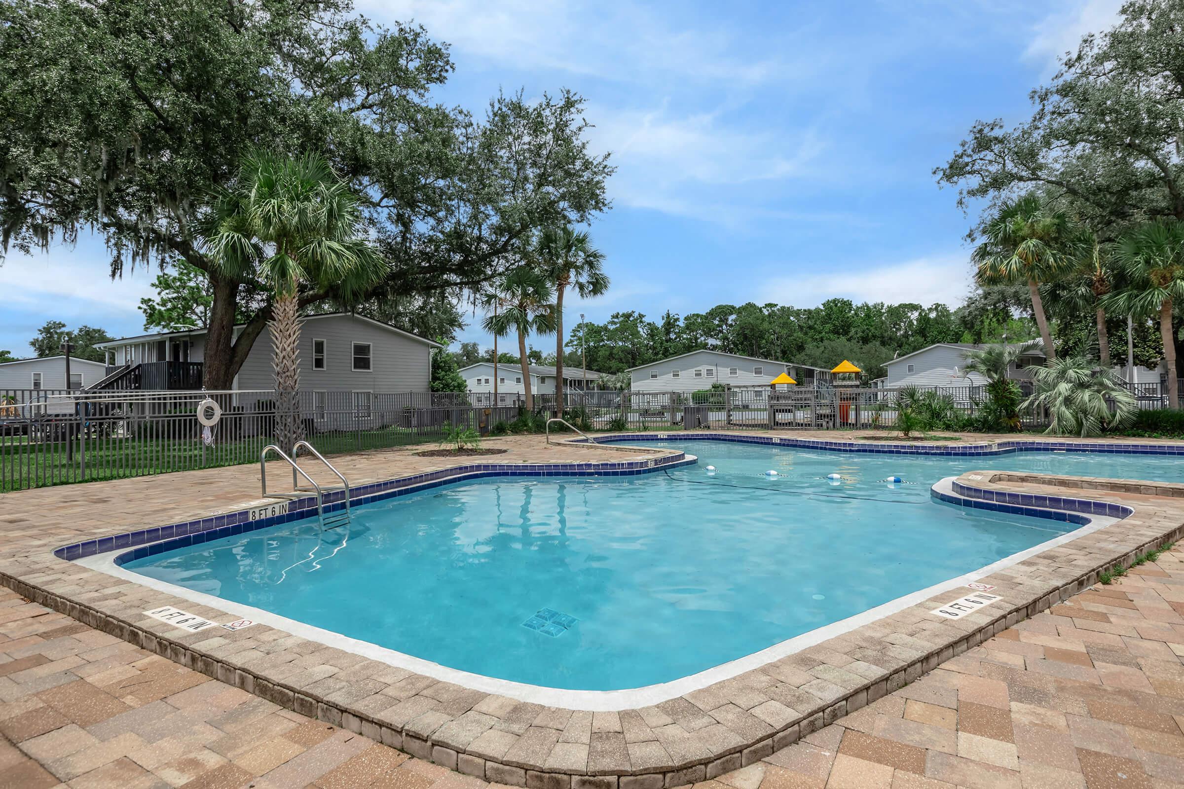 A clear swimming pool with a light blue surface, surrounded by a patio made of square tiles. Lush green trees and palms provide shade around the area, while nearby, there are various residential buildings and recreational structures in the background under a blue sky.