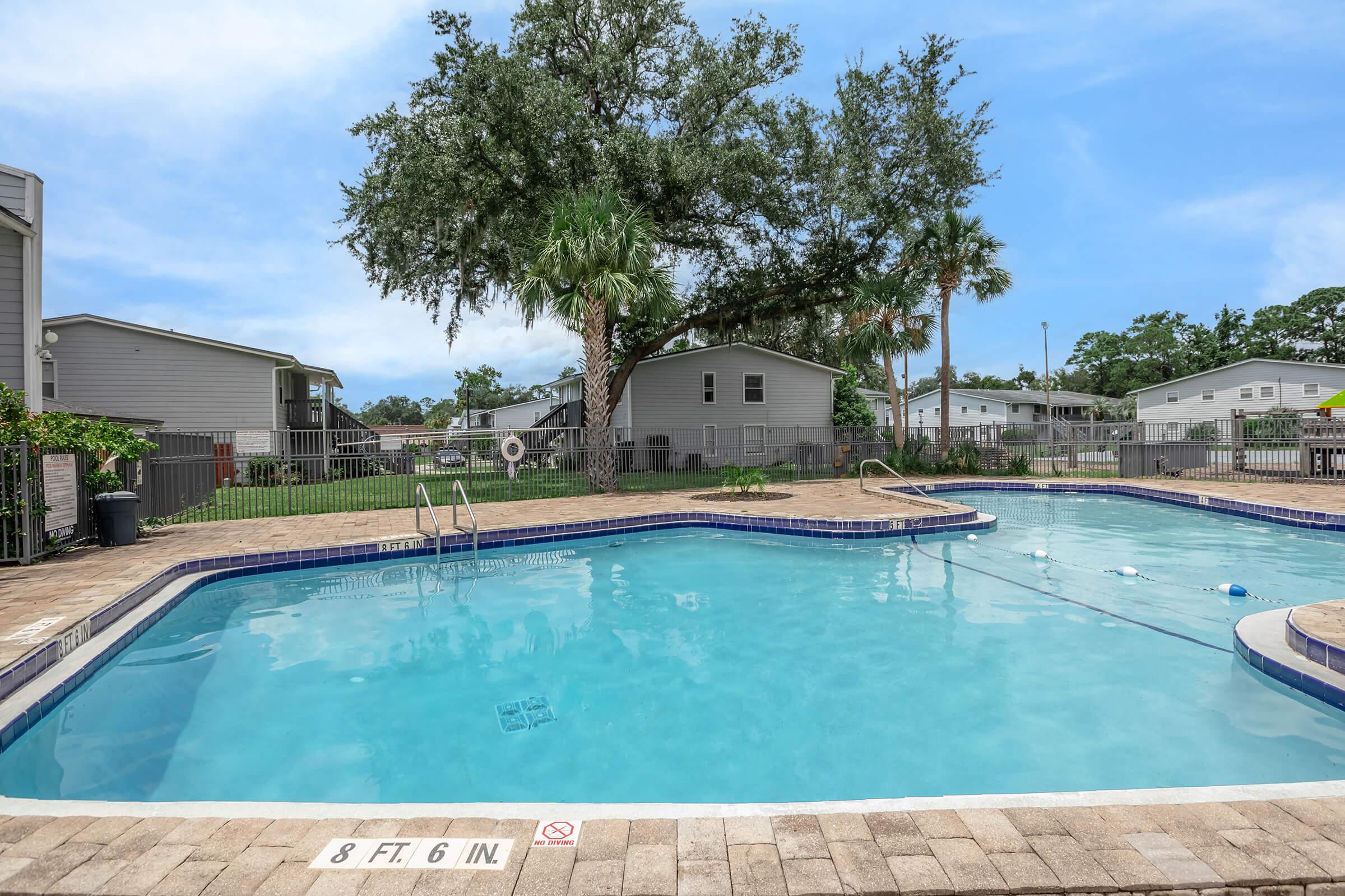 Swimming pool surrounded by paved patio and palm trees in a residential area. In the background, there are several homes and a green lawn. A depth marker indicates the pool's depth of 8 feet 6 inches. Clear blue sky overhead creates a bright atmosphere.