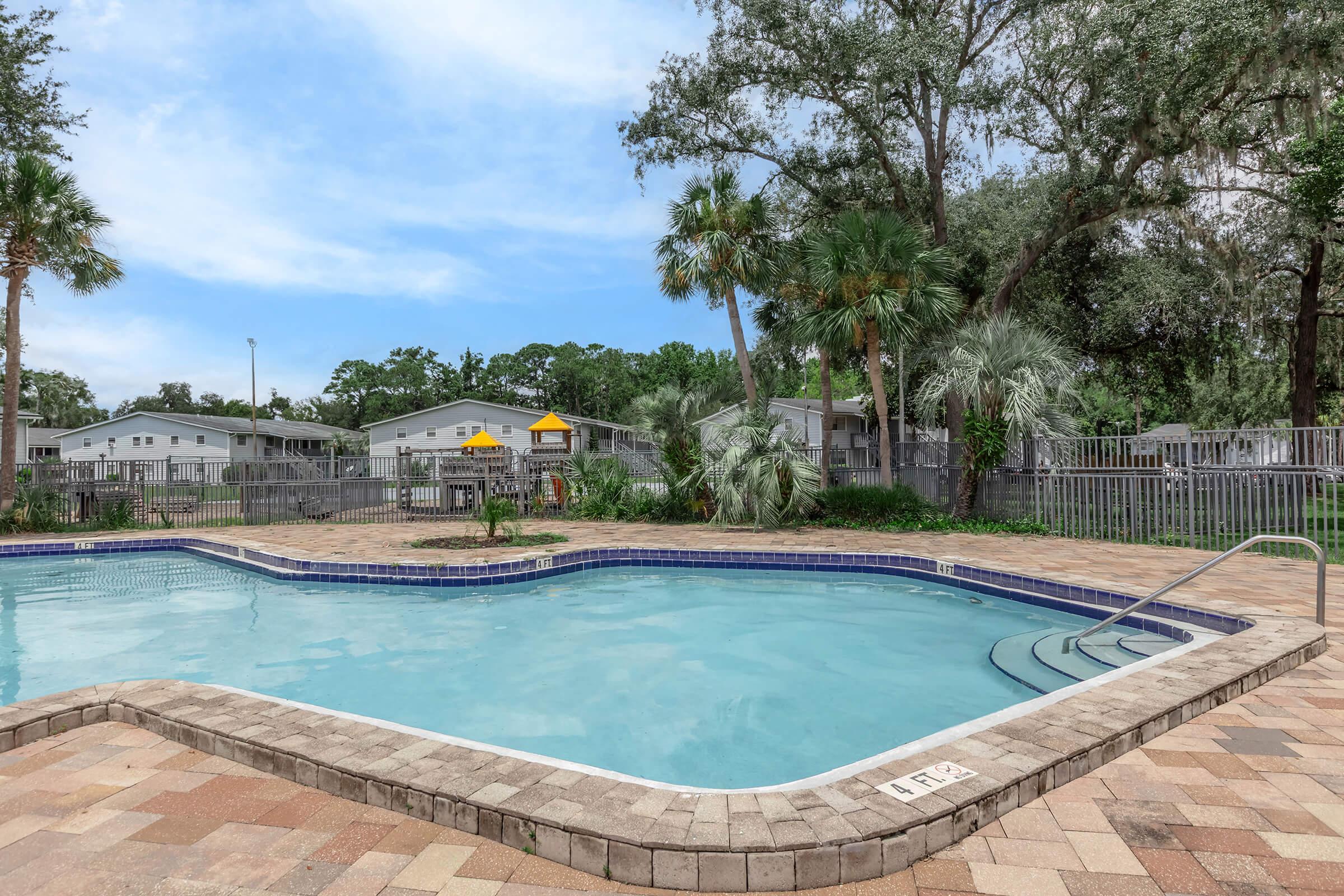 A clear swimming pool with a rounded edge surrounded by a stone patio. Lush green palm trees and shrubs line the area, with residential buildings in the background. The sky is partly cloudy, and a sunny atmosphere is evident. A small staircase leads into the pool.