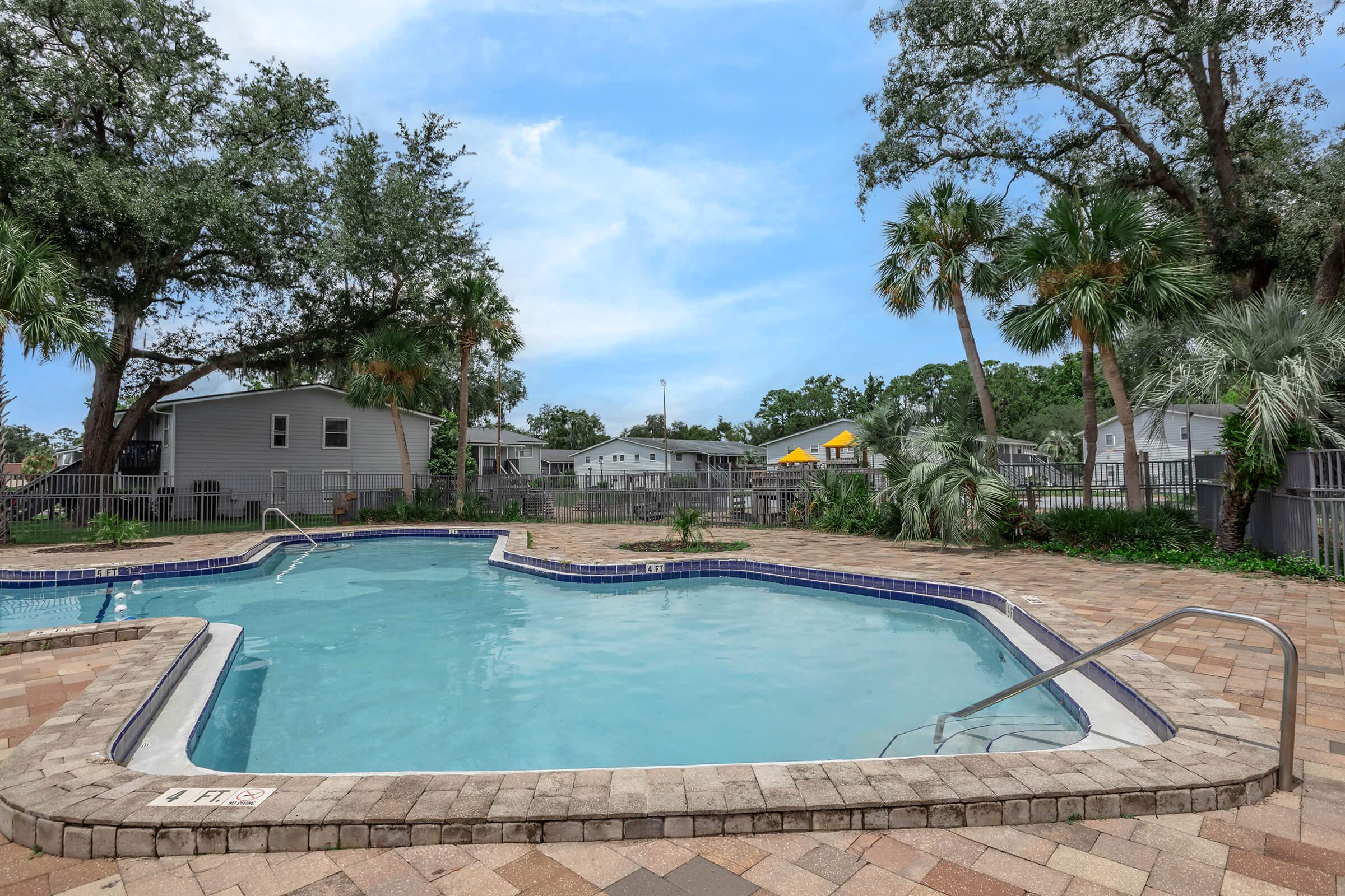 A serene outdoor pool area with clear blue water, surrounded by palm trees and landscaped pathways. Nearby, there are residential buildings, some with yellow accents, under a bright blue sky with scattered clouds. The patio features textured stone flooring, creating a welcoming atmosphere for relaxation.